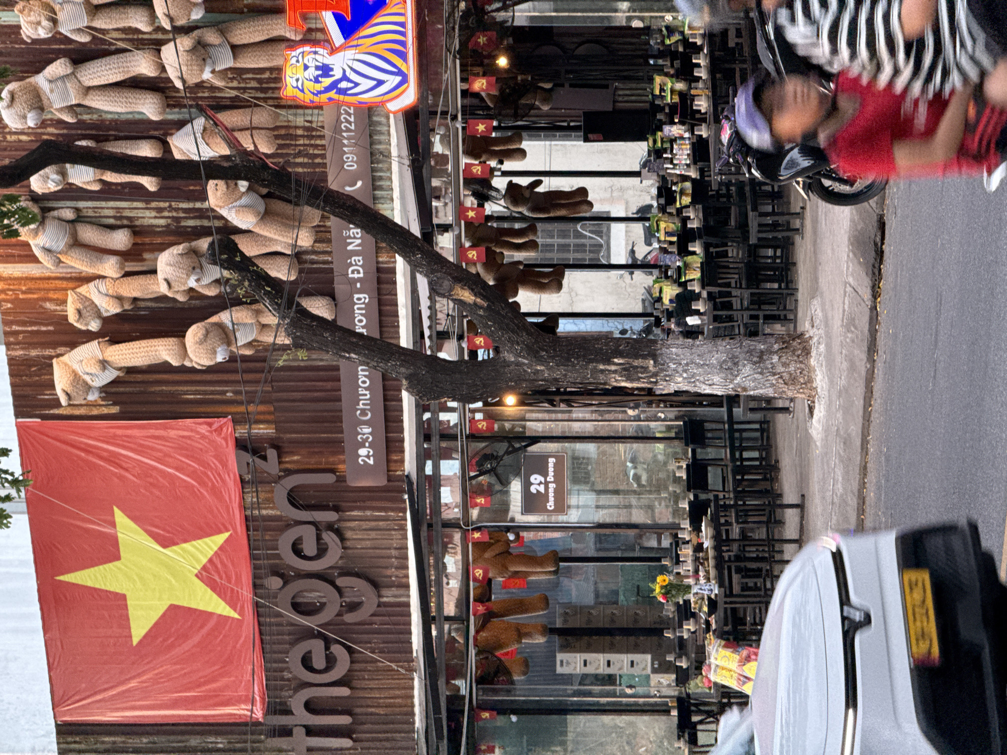 The image captures a street scene in Da Nang, Vietnam. The facade of a building, adorned with a large red flag featuring a yellow star (likely the Vietnamese national flag) and several plush teddy bears, is prominent. Below the flag, the building's signage reads "the gen²". A street number "29-30 Chuong Duong" is visible on a brown sign. A blue and yellow neon sign depicting a tiger is also present. The street in front of the building is lined with tables and chairs, suggesting an outdoor seating area for a restaurant or cafe. A white car is partially visible in the foreground, with a yellow license plate. A motorbike carrying two people, one wearing a red shirt and a purple cap, is moving on the street. The environment appears to be urban, with trees and a paved road. The overall impression is a lively street-side establishment in Vietnam.