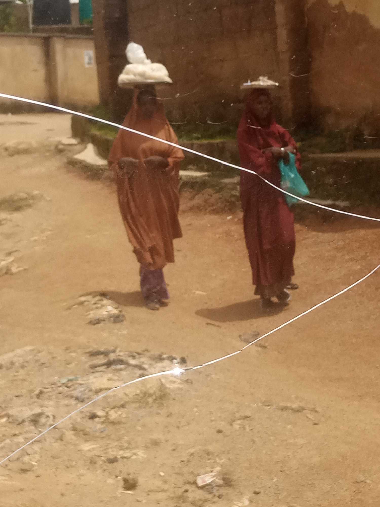 Two women are walking on a dirt road in Jos, Nigeria. The woman on the left is wearing a brown abaya and a headscarf, carrying a tray of white items on her head. She is also wearing purple trousers. The woman on the right is wearing a maroon abaya and a headscarf, and is carrying a blue bag in her left hand. Both women are wearing sandals. The ground is dusty and there are some scattered pieces of debris. In the background, there are some low-rise buildings. The image is slightly blurry and has some white lines across it, suggesting it might be a photograph taken through a dirty or cracked window.
