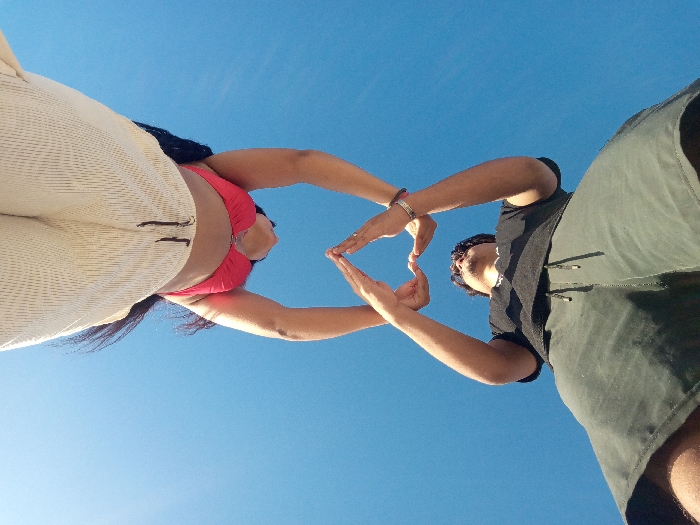 Two people pose against a clear blue sky. One person wears a red top and light-colored pants, with their dark hair visible. The other person is in a dark t-shirt and green shorts. They both hold up their hands, forming a heart shape with their fingers in the open space between them. The image is taken from a low angle, emphasizing the sky and the subjects above.