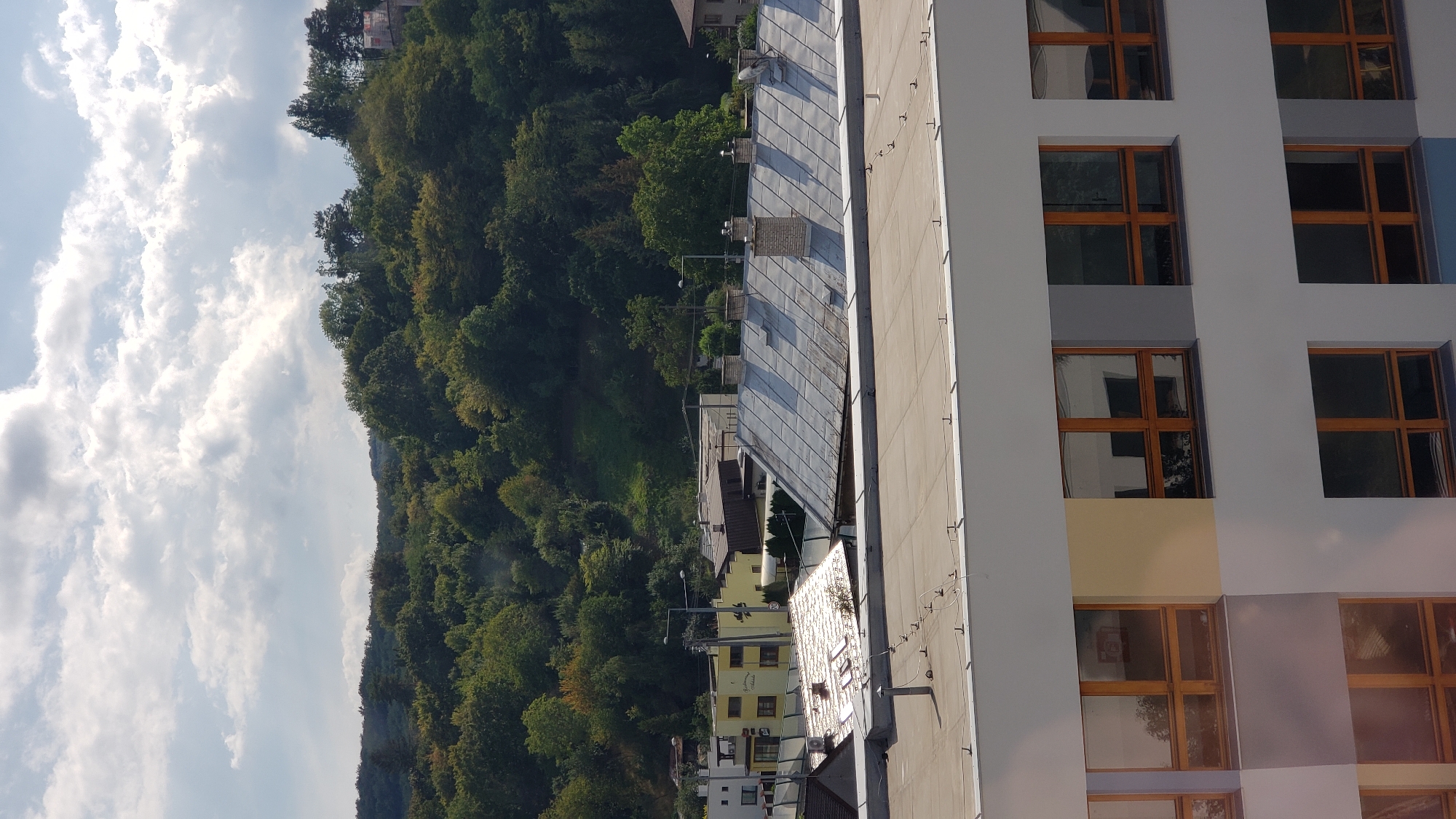 This is an eye-level, medium-range, vertical shot of a suburban landscape in Prešov, Slovakia. The image is dominated by a large, modern building in the foreground with large, rectangular windows with wooden frames. Behind this building, there are several rooftops of older, smaller houses with tiled roofs and chimneys. Further in the background, a lush, green, forested hillside rises, with scattered houses and trees visible amongst the foliage. The sky above is partly cloudy, with patches of blue sky showing through. The overall impression is one of a transition between urban and natural environments.