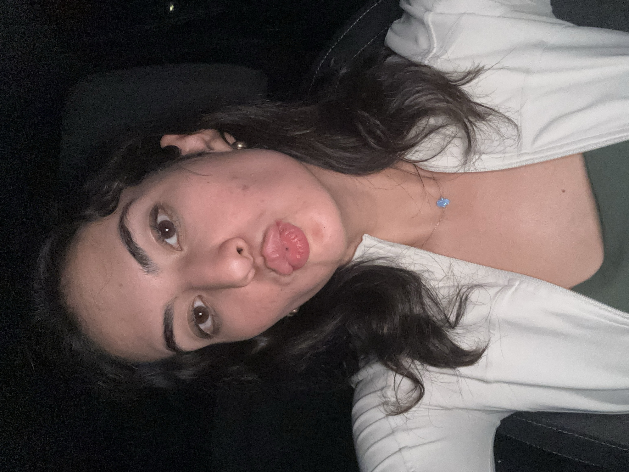 A young woman with dark hair and brown eyes is making a kissing face at the camera. She is wearing a white jacket and a green top. She is also wearing a necklace with a blue pendant. The background is dark. The location is uncertain and cannot be determined from the visual information provided.