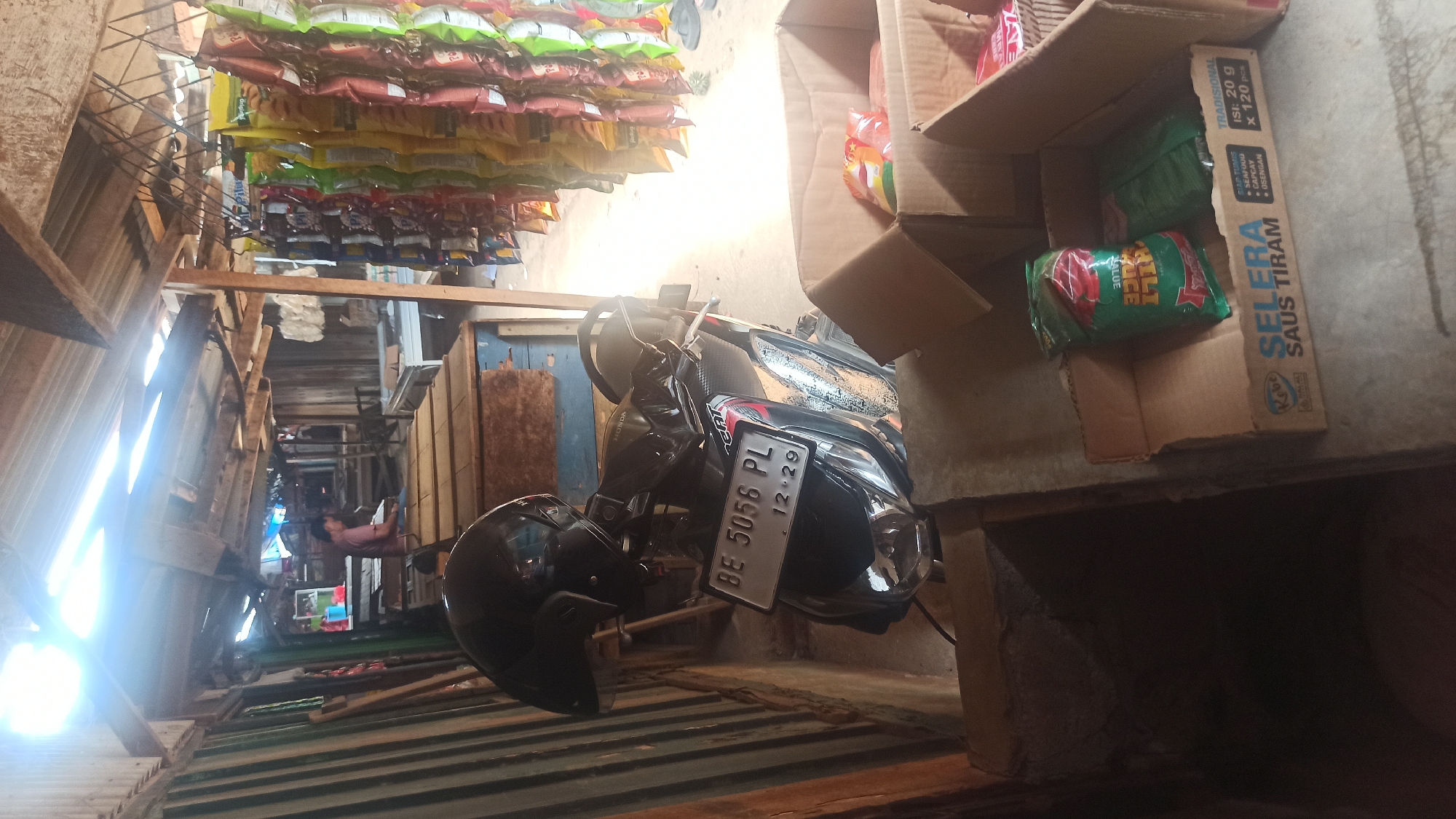 In a dimly lit, open-air market stall in Trisinar, Indonesia, a black motorcycle with license plate "BE 5056 PL" is parked. A black helmet rests on the handlebars. The stall features a corrugated tin roof supported by wooden beams, with various goods displayed. To the right, rows of packaged snacks hang from a wire rack. In the background, a person is seated at a wooden counter. In the foreground, two cardboard boxes are placed on the ground. The left box contains several small packages of food, and the right box also holds food items, with one package prominently displaying "SELERA SAUS TIRAM" in Indonesian. The environment appears rustic and functional, typical of a local market.