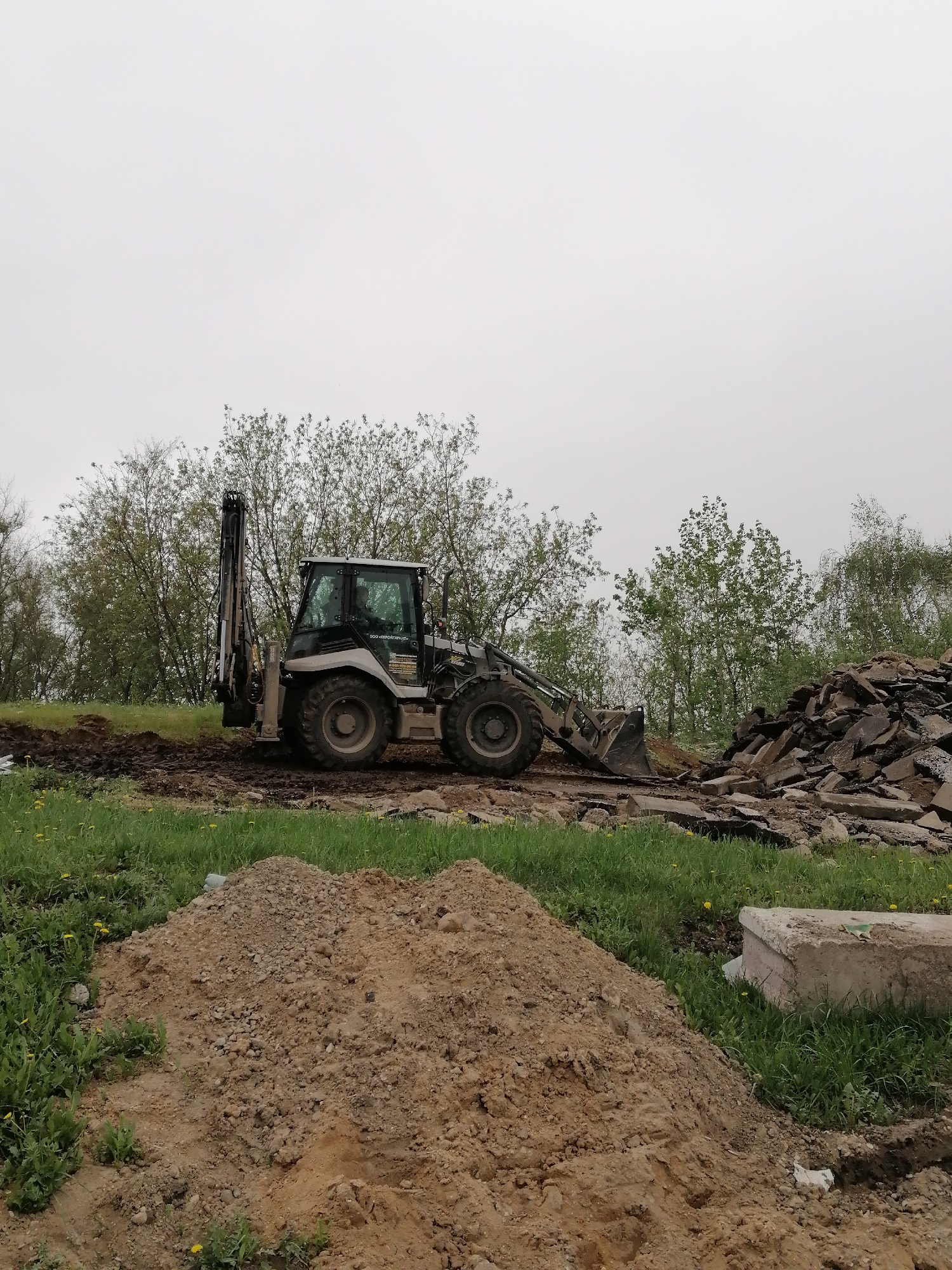 The image depicts a construction or demolition site, possibly located in the Moscow region of Russia, indicated by the Cyrillic script on the construction vehicle. A backhoe loader sits in the middle ground, positioned between a pile of broken asphalt or concrete debris and a mound of dirt. Green grass and scattered dandelions surround the disturbed earth. Trees form a backdrop against an overcast sky. No people are visible.
