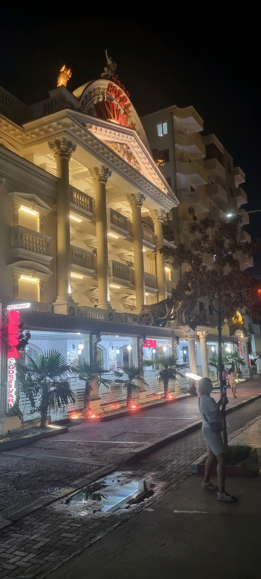 The image shows the facade of a multi-story building at night, likely a hotel or restaurant, in Bashkia Durrës, Albania. The building features classical architectural elements, including prominent columns, balconies with ornate railings, and statues. A large, illuminated sign with red lettering that appears to say "HOTEL" is visible on the upper facade, along with statues of figures and a golden hand. The lower level of the building has a row of windows and a covered walkway with palm trees and decorative lighting. A neon sign reading "RESTORANT" is mounted vertically on the left side of the building.
In the foreground, a paved sidewalk and street area are visible. A puddle of water on the street reflects the lights from the building. A woman in a light-colored top and shorts, with her back to the camera, is standing on the sidewalk, holding onto a tree trunk. In the background, a residential building with multiple floors and balconies is visible to the right of the main structure. The scene is illuminated by artificial lighting, suggesting it is nighttime.