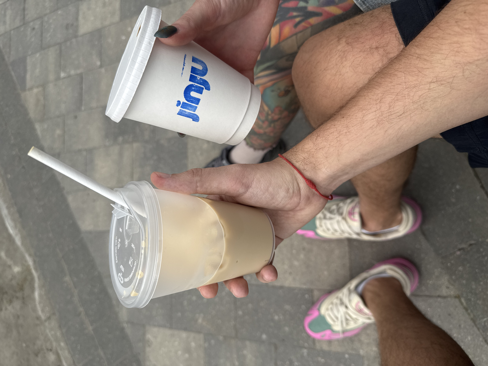 A medium-shot photograph shows two beverages being held outdoors over gray stone pavers. One beverage, a paper cup labeled "Jinju" in blue, is in a person's left hand; the person has dark nail polish on at least one finger. The other beverage is in a clear plastic cup with a straw, filled with a light brown liquid, held in another person's right hand. The legs and feet of the two individuals are visible in the frame, revealing a pair of athletic shoes with white laces and pink and green detailing on the lower subject, and a glimpse of another shoe on the upper subject. The upper subject has tattoos on their leg and is wearing dark shorts. The lower subject is wearing socks and has a red wristband. No other identifying features or information is visible in the photograph. There is no definitive information to prove this image was taken in Moscow, Russia.