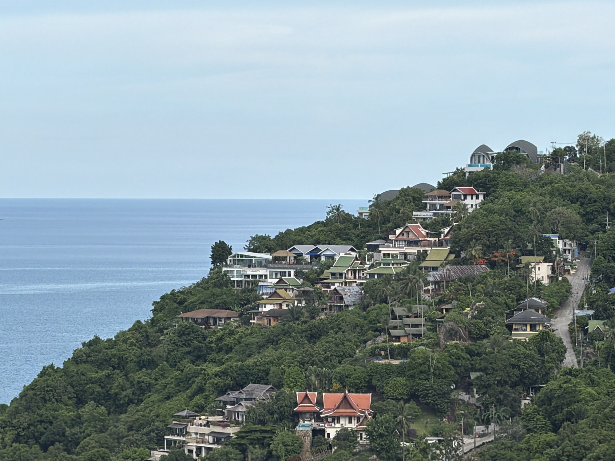 A wide-angle, high-angle, daytime shot captures a hillside village in Baan Chaweng Noi, Thailand, overlooking the ocean. The densely forested slope is dotted with numerous buildings, varying in size and architectural style, with many featuring red-tiled roofs. The ocean, a calm, deep blue, stretches to the horizon under a pale blue sky with wispy clouds. A road winds its way up the hill, bordered by lush green trees and vegetation. Several houses are visible, some with balconies and private pools. The scene suggests a residential area with a focus on integration with the natural landscape.