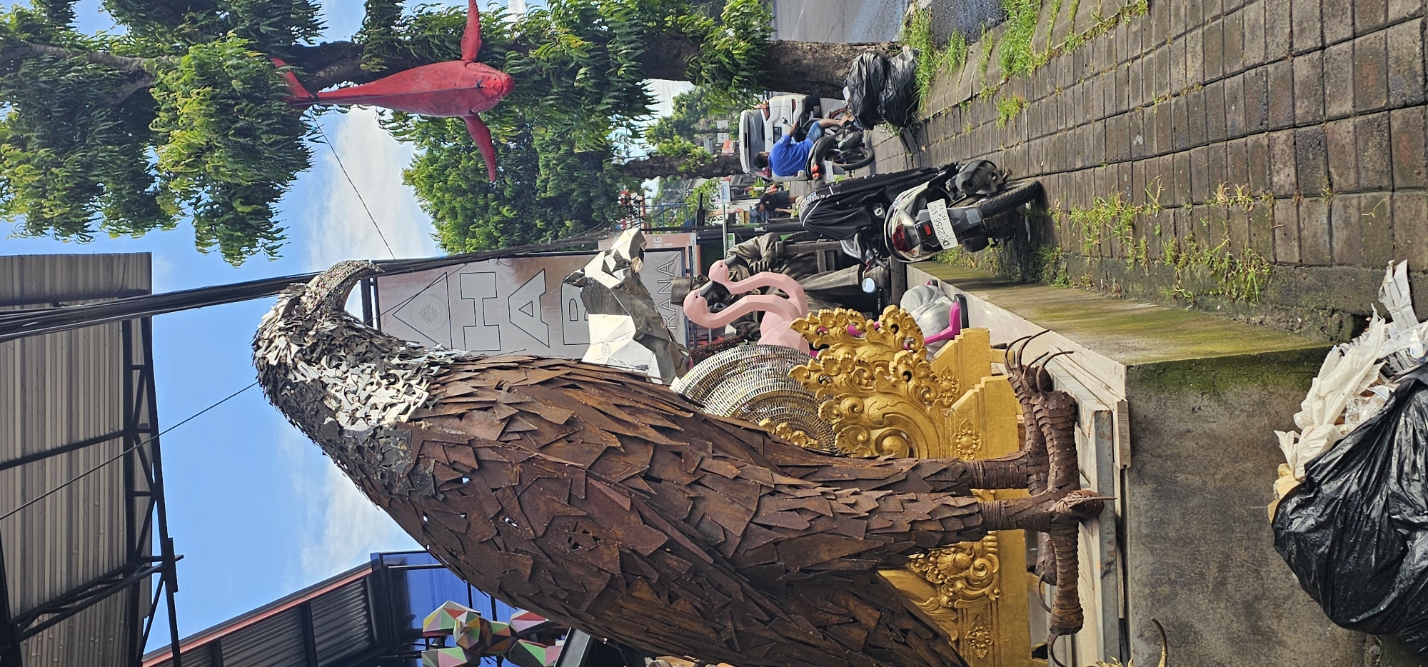 The image captures a scene in what appears to be the Ubud District of Indonesia. In the foreground, a large, elaborate sculpture of an eagle crafted from pieces of metal dominates the view. Behind the eagle are various other decorative objects including pink flamingos and ornate gold-colored structures.
In the background, a street scene unfolds, with parked motorbikes along a paved sidewalk. A person in a blue shirt is visible sitting on one of the motorbikes. A red, fish-shaped decoration is suspended from a tree. The environment consists of lush greenery from the trees and a cloudy sky. The overall impression is that of a vibrant, artistic street scene in a busy urban environment.