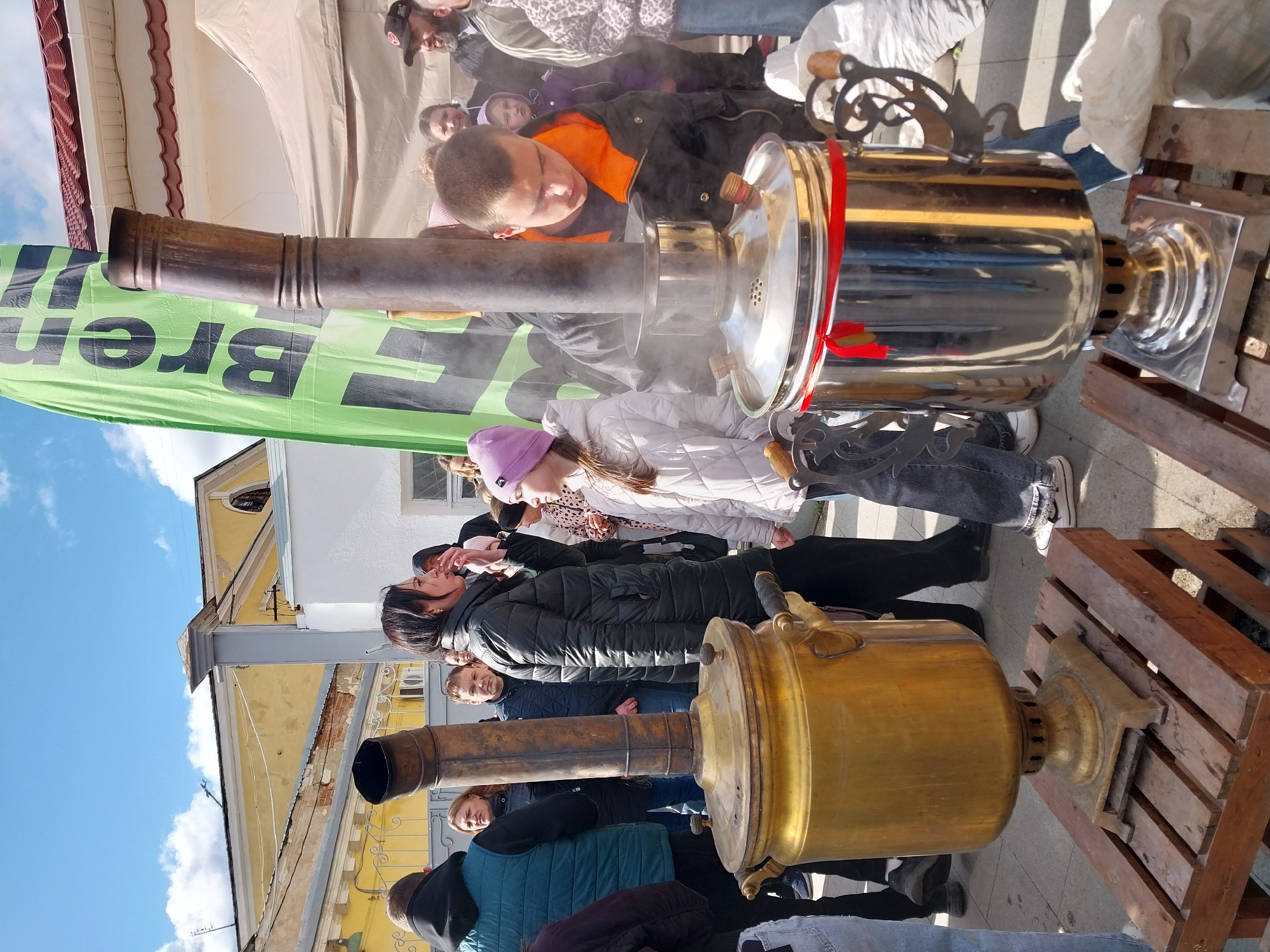 The image depicts a scene in Orenburg, Russia, showing two large, traditional Russian samovars (tea urns) set up outdoors. One samovar is golden in color with a prominent chimney, while the other is silver with a red ribbon. A group of people of various ages are gathered around the samovars. A green banner with black lettering is visible behind one of the samovars. The background includes buildings with white and yellow facades, and a patch of blue sky with clouds is visible above. The setting appears to be a street or public area.