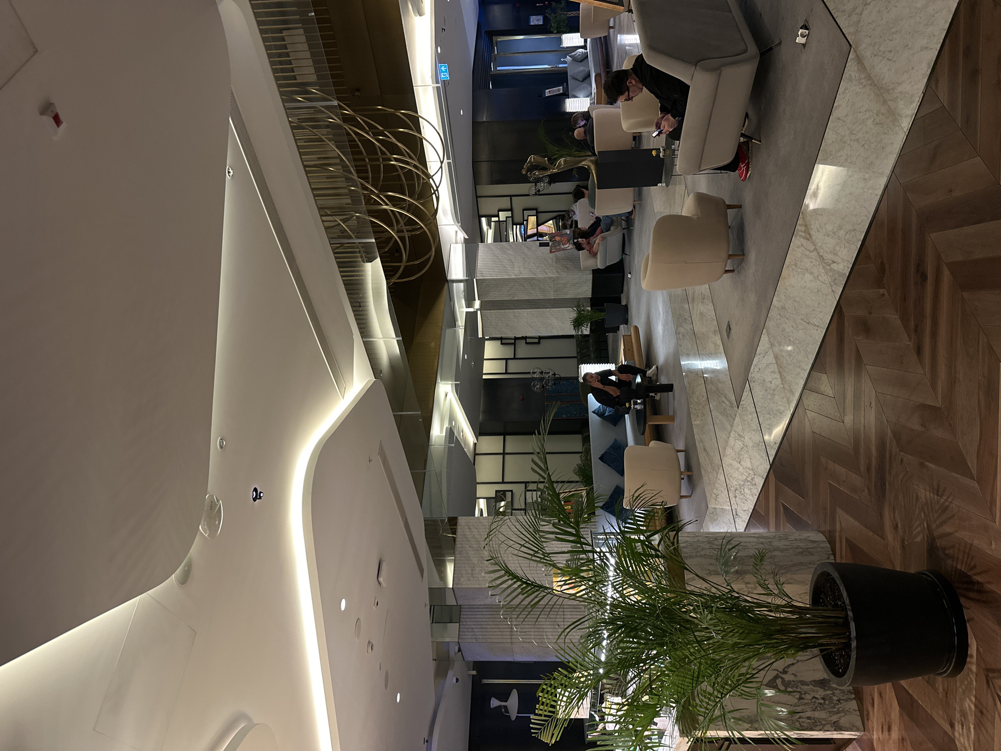The image depicts the lobby of a building in Batumi, Georgia. The space is modern and features a combination of architectural elements including white curving ceilings, brass decorative rings, and vertical striped walls. People are scattered throughout the lobby, some seated in chairs and sofas, apparently engaged in individual activities such as using mobile phones or reading. The flooring is comprised of alternating sections of marble and herringbone-patterned wood. Large potted plants are placed within the seating areas, contributing to the interior decor.