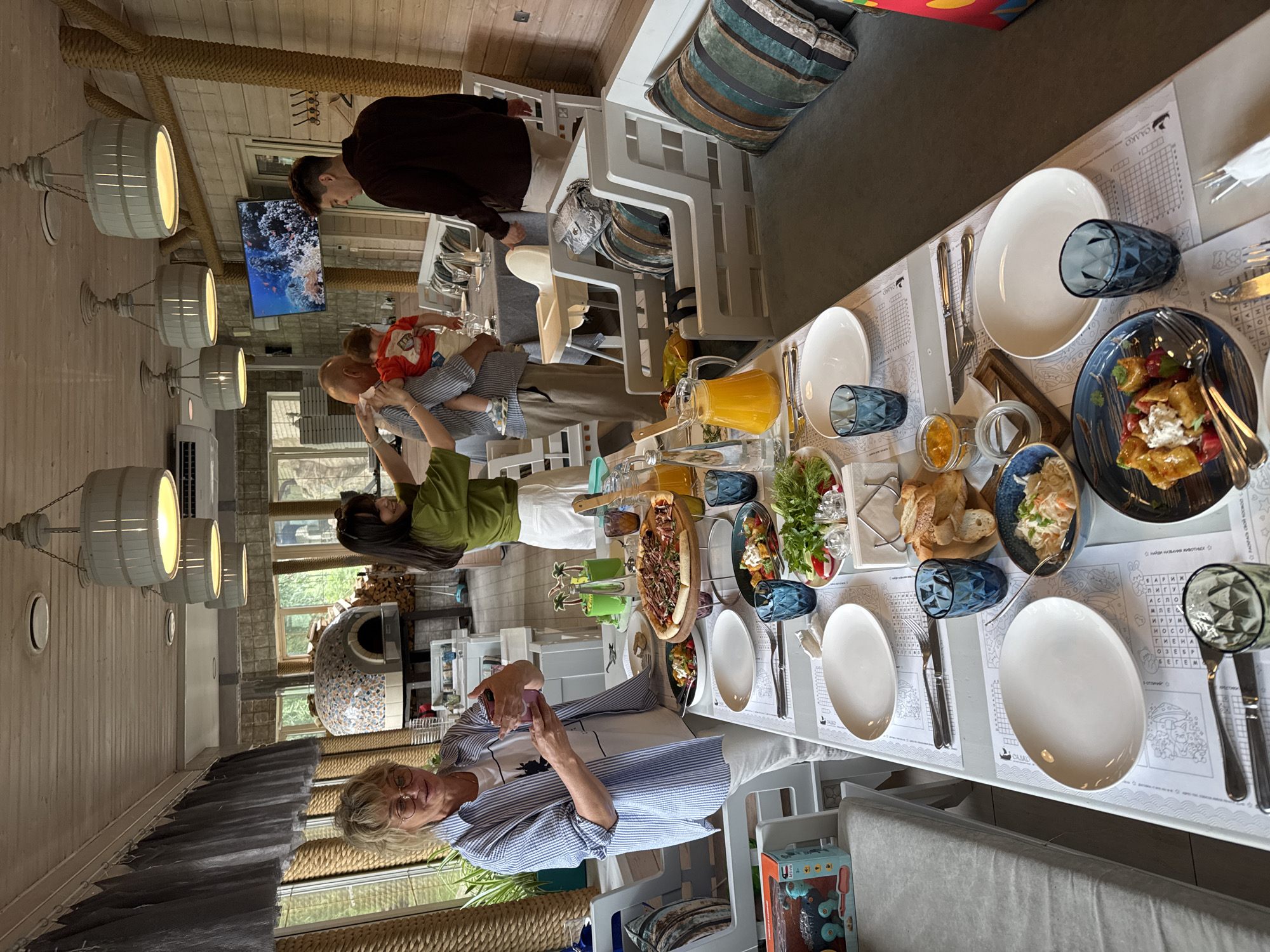 A group of people are gathered around a table in a restaurant. The table is set with plates, glasses, and various dishes, including pizza, salad, and what appears to be a fruit compote. A woman in a blue and white striped shirt is holding a drink, and a man in a dark shirt is standing in the background. A child is being held by an adult, and other people are seated at the table. The restaurant has a rustic interior with wooden walls and ceiling, and there are several pendant lights hanging overhead. A pizza oven is visible in the background. The scene appears to be a family gathering or celebration.