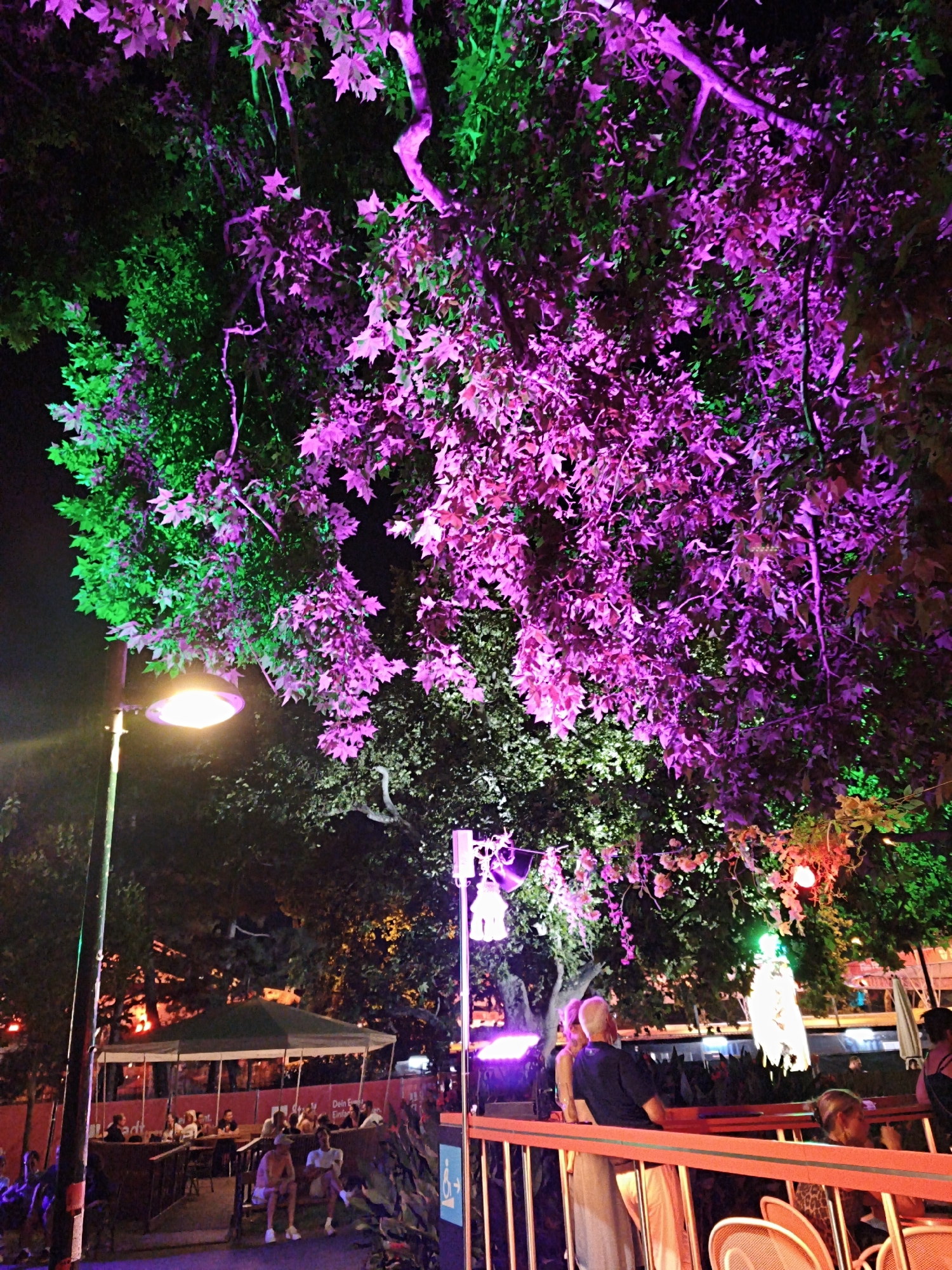The image captures an outdoor scene at night, likely a public gathering or event space in Vienna, Austria. Dominating the upper portion of the frame are the branches and leaves of large trees illuminated by vibrant green and purple lights, creating a striking, artificial glow against the dark sky. In the middle ground, a street lamp with a bright, warm light is visible on the left, casting illumination on its pole and the surrounding area. Further back, beneath the illuminated trees, a seating area is set up with tables and chairs, some covered by a large tent or canopy. Several people are seated at the tables, engaged in conversation or simply enjoying the ambiance. On the right side of the image, a bright orange railing runs across, with several people standing or sitting behind it. One individual in the foreground is seated on a bench, facing away from the camera. In the background, more trees and structures are faintly visible, also lit by ambient lighting. The overall atmosphere suggests a lively, festive evening event.