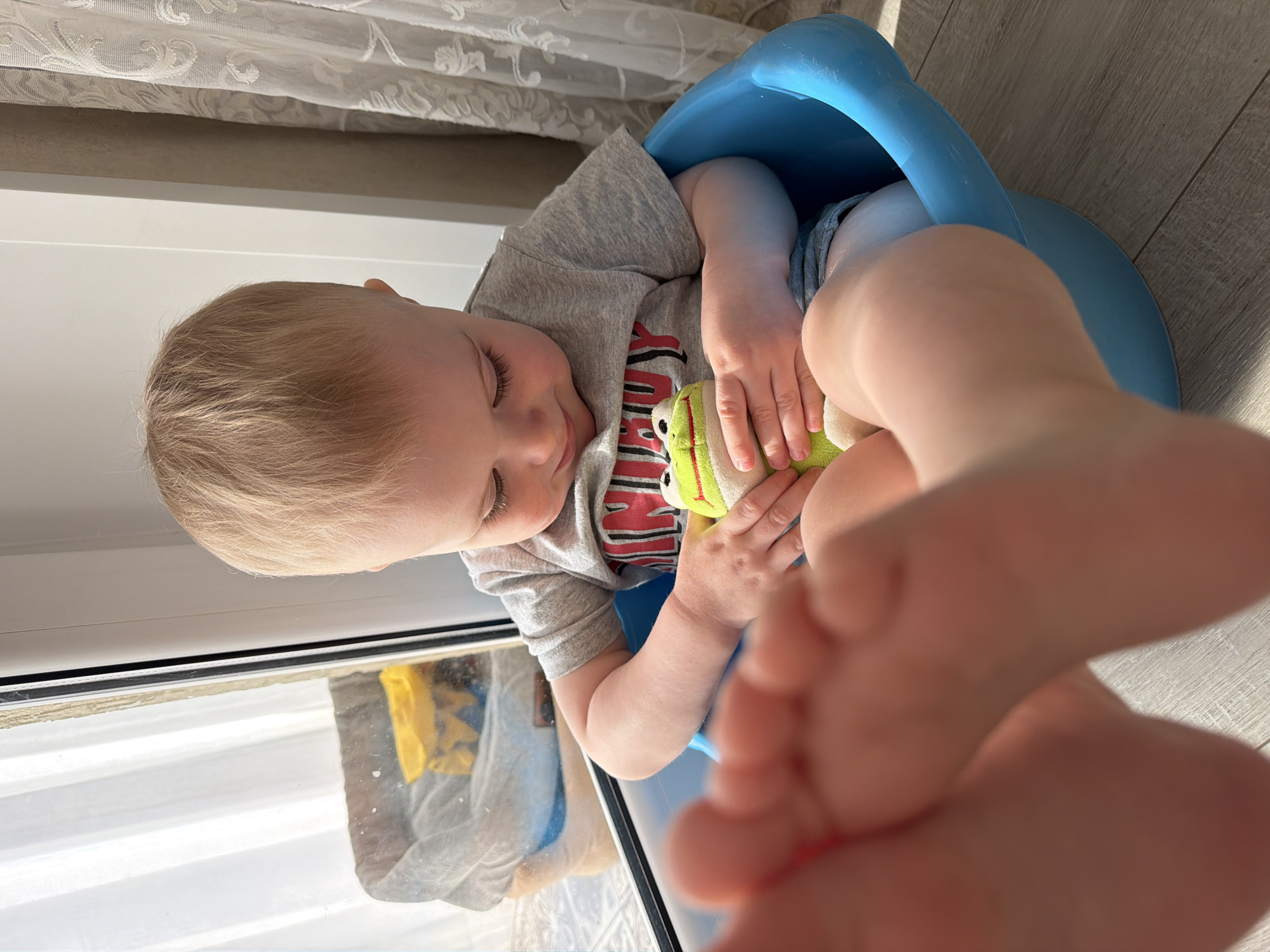 A young child is sitting on a blue potty. The child has blonde hair and is wearing a grey t-shirt with a red and black design. The child is holding a green and yellow stuffed toy. The child's foot is in the foreground, partially obscuring the view. A window is to the left, reflecting what appears to be a curtain and possibly another object. The location is unknown but purported to be Votkinsk, Russia. The setting appears to be indoors, possibly a residence.