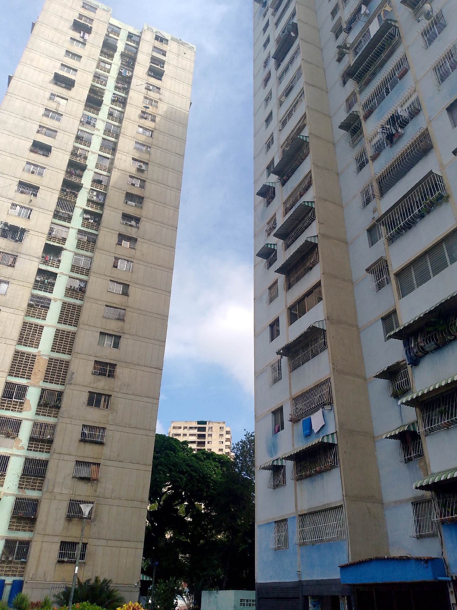 The image depicts two tall residential buildings in Caracas, Venezuela, angled upwards towards a partly cloudy sky. The building on the left is primarily beige in color with numerous windows and some balconies. The building on the right is painted blue, also featuring many windows, balconies, and some window awnings. Clothes are hanging out to dry on some of the balconies. Below the buildings, there are trees and smaller buildings.