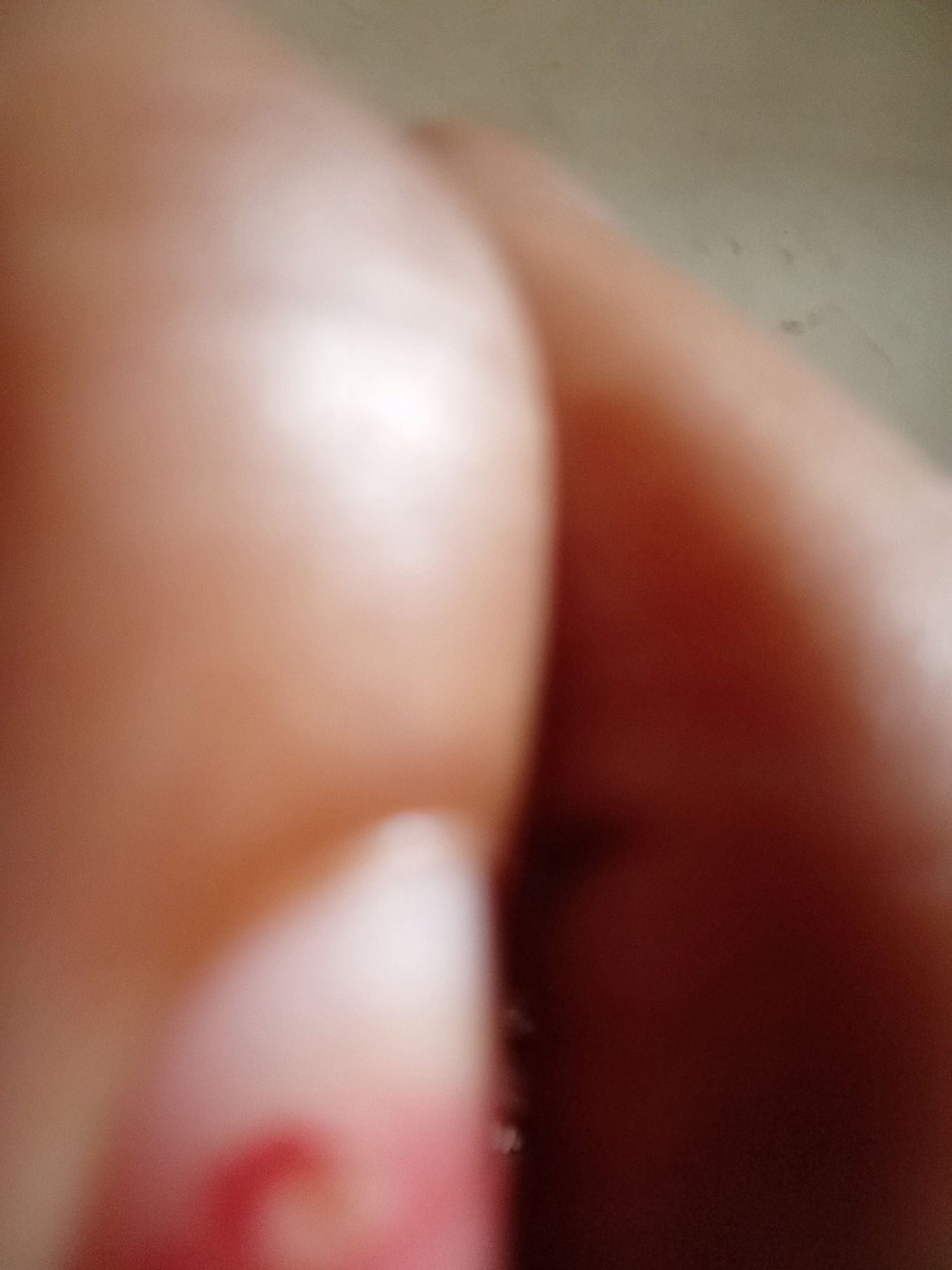This is a close-up, out-of-focus media file. The primary subjects appear to be human skin, possibly from a hand or arm, due to the smooth texture and pinkish-brown tones. The lighting suggests an indoor setting, with a bright light source creating highlights on the skin. There is a hint of red color in the lower left quadrant, which could be a nail polish or a mark on the skin. The background is indistinct and appears to be a muted, neutral color. The overall impression is abstract and lacks clear detail due to the lack of focus. No text or discernible activity is visible. The time of day and weather are not inferable.