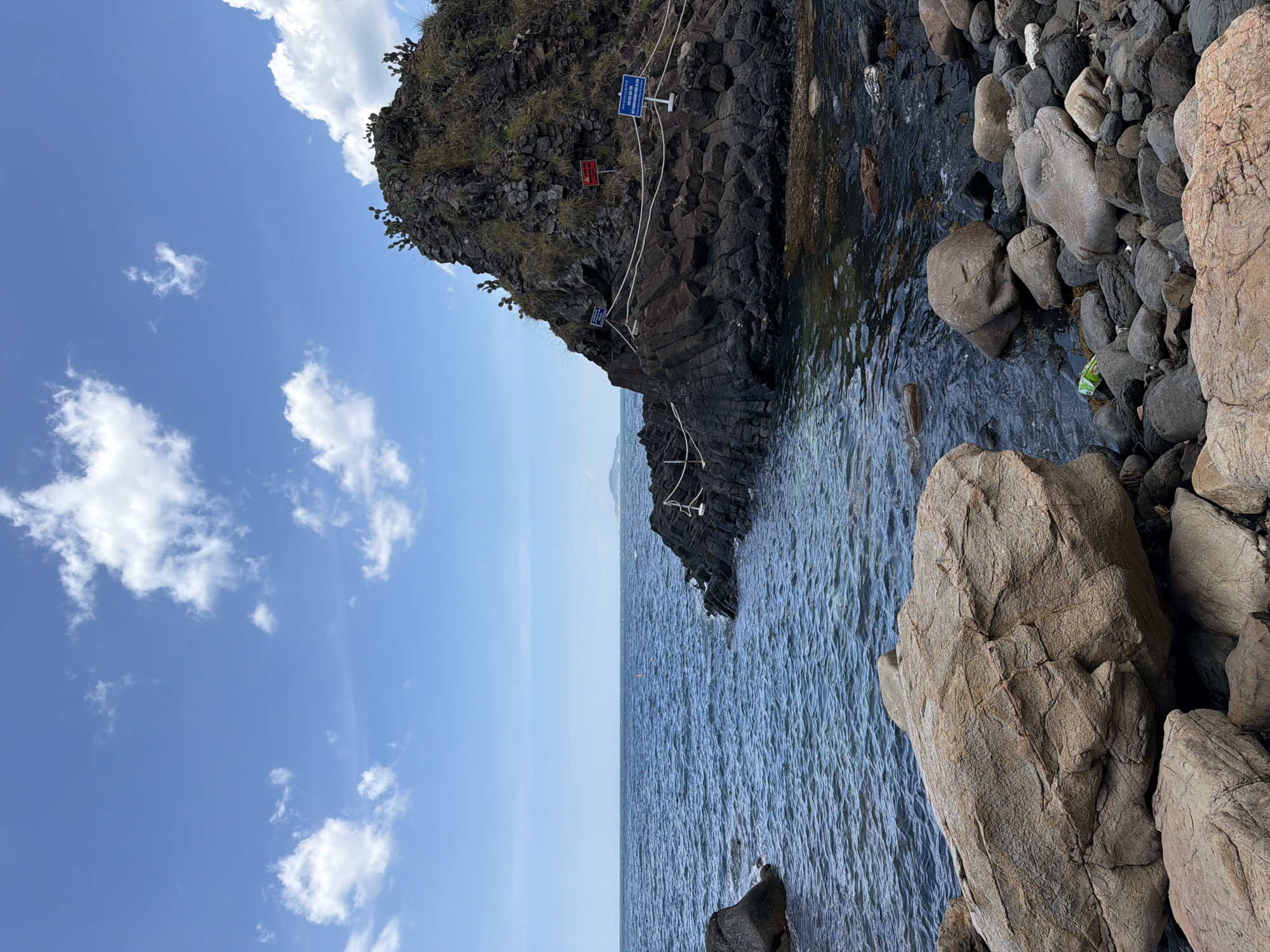 The image depicts a rocky shoreline and a dark volcanic rock formation jutting out into the ocean under a bright blue sky with scattered white clouds. The water is a deep blue with ripples. Several large, light-brown boulders are in the foreground, with smaller dark rocks and pebbles leading down to the water. On the volcanic rock formation, there are signs and a rope barrier, suggesting it is a designated area or a point of interest. Sparse green vegetation, possibly cacti, grows on the top of the formation. The scene is located in Phú Lương, Vietnam, and appears to be a natural landscape with some minor human intervention for visitor management. There are no people visible in the image.