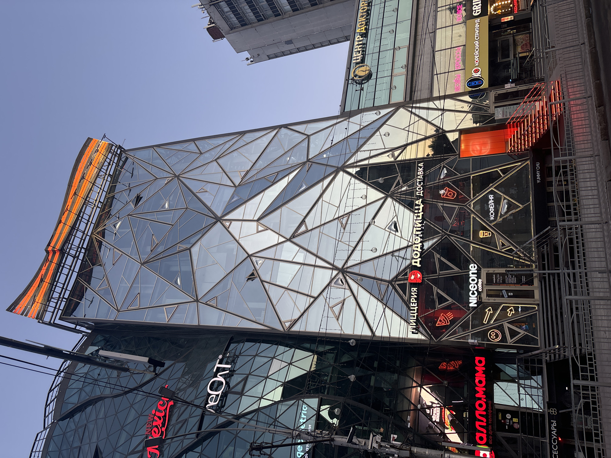 This is a daytime, eye-level exterior shot of a modern building complex in Novosibirsk, Russia. The building features a distinctive, geometric glass facade with a multifaceted, angular design that reflects the sky and surrounding structures. The structure has multiple levels with businesses located at the ground floor, accessible by stairs and entrances. Prominent signage for various establishments is visible, including "New texter," "eOT," "Alllo mama," "Pizzeria Dodo Pizza Delivery," "Niceone coffee," "Yummy Chu," and "Center Doktorov." The building is illuminated by neon and LED signs, some of which are red and orange, creating a vibrant contrast against the glass and sky. There are visible power lines and streetlights in the foreground, suggesting an urban environment. The sky is clear and blue, indicating fair weather. No people are clearly visible in the image.