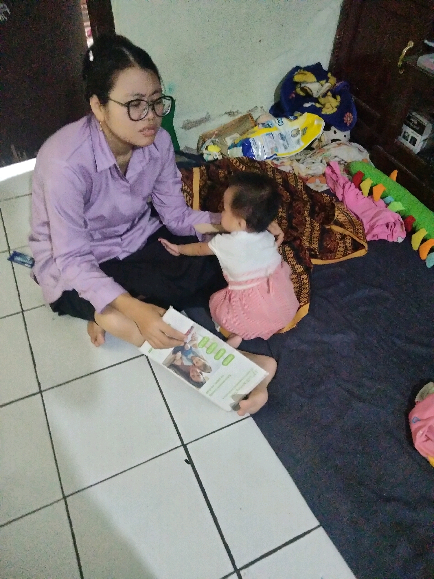 In Suka Sari, Indonesia, a young woman wearing glasses and a purple shirt sits on a tiled floor, holding a book. She is facing a baby who is sitting on a dark blue blanket, wearing a pink dress. The baby is reaching towards the book. Behind them, a pile of blankets and toys, including a green stuffed animal, are scattered on the floor and on a wooden shelf. The room appears to be a living area.
