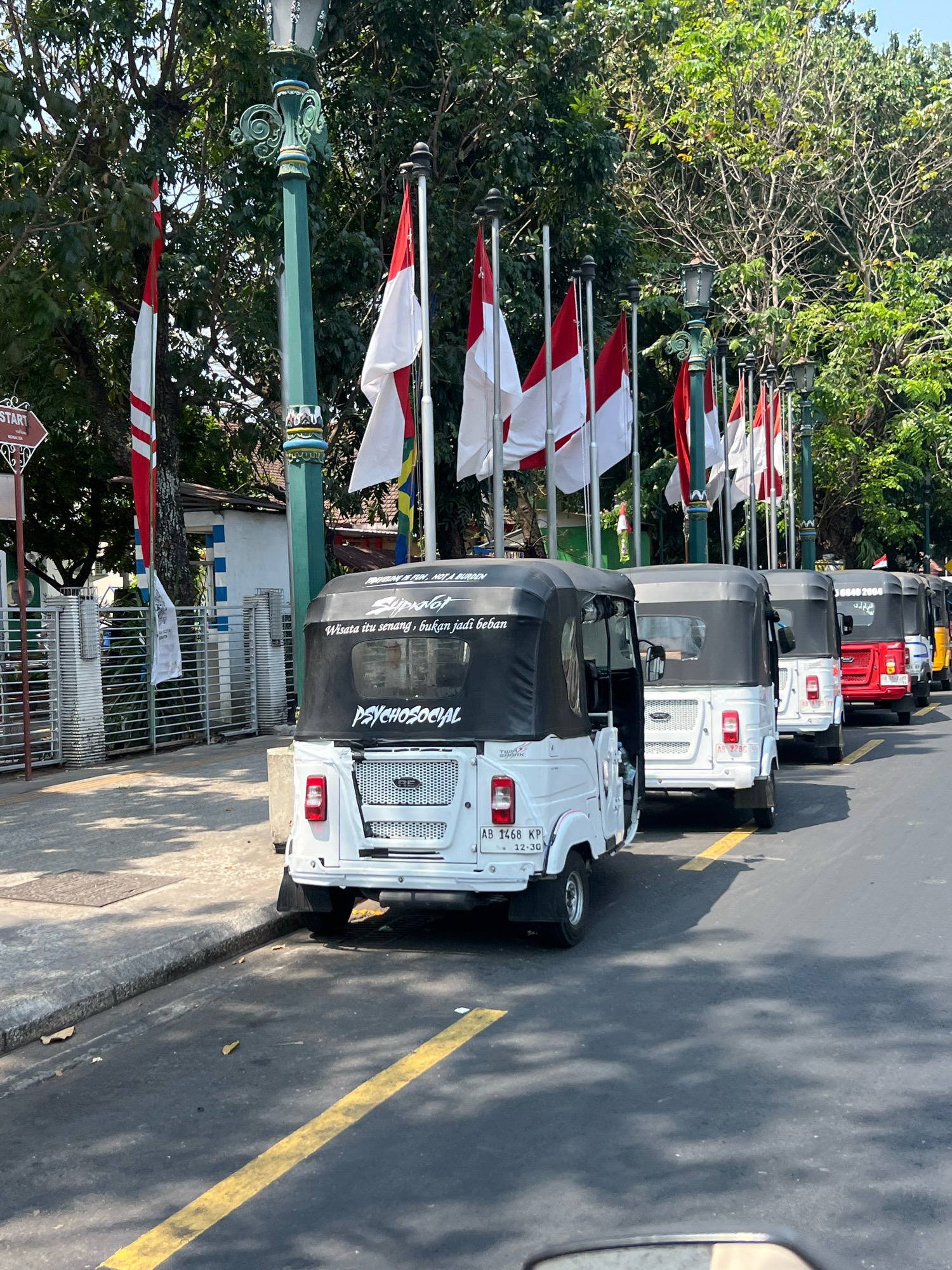 A line of white and red three-wheeled vehicles, commonly known as "bajaj" or "betor," are parked along a street in Yogyakarta, Indonesia, with lush green trees and Indonesian flags lining the sidewalk. The bajaj in the foreground has "Slipknot" and "Wisata itu senang, bukan jadi beban" (Tourism is fun, not a burden) written on its black canopy, along with "PSYCHOSOCIAL" in white lettering. The vehicles are positioned on a paved road with a yellow painted line marking a lane. In the background, a decorative lamppost stands near a fence and a small building. The scene appears to be a sunny day, with shadows cast on the road.