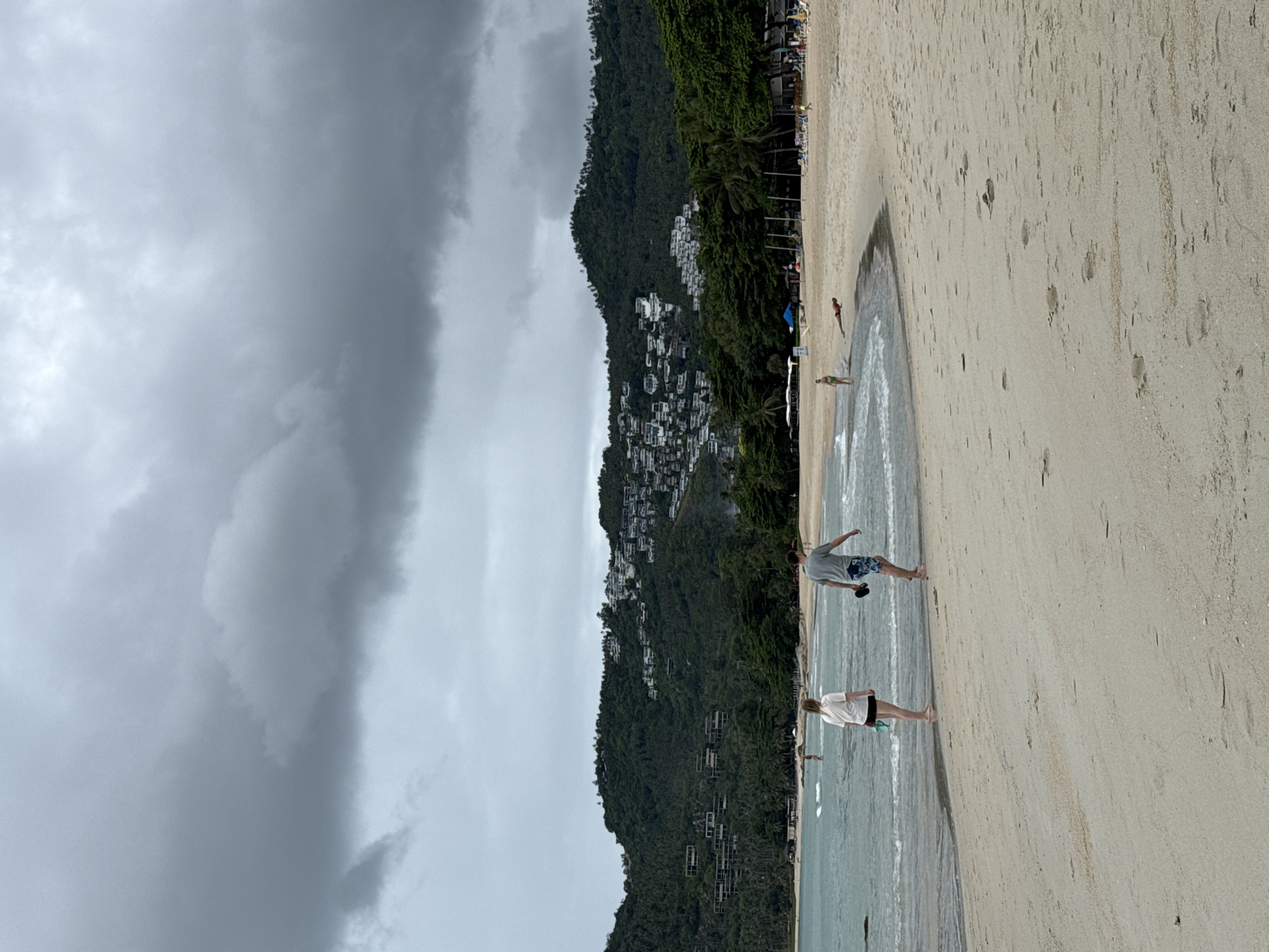This is an eye-level, medium shot of a beach on a cloudy day in Koh Samui District, Thailand. In the foreground, two people are walking on the sand near the water's edge. A man in a grey t-shirt and patterned shorts walks away from the camera, holding a pair of flip-flops. Beside him, a woman in a white t-shirt and dark shorts walks towards the camera, also holding flip-flops. The water is calm with small waves lapping at the shore. In the background, a densely forested hillside is covered with white buildings, indicating a resort area. The sky is overcast with dark, heavy clouds. Beach chairs and umbrellas are visible further down the beach.