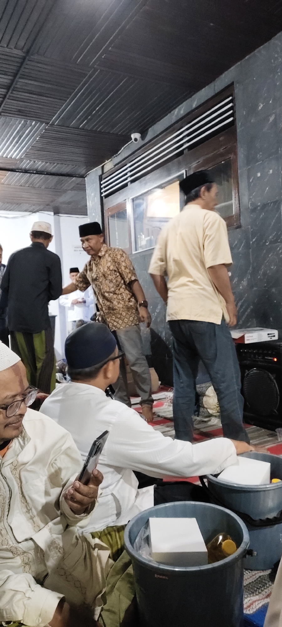In a dimly lit interior space, possibly a hall or meeting room, several men are gathered. The ceiling is covered with dark, striped panels. A security camera is visible on the ceiling near a window with horizontal blinds. In the foreground, a man wearing glasses and a white peci (cap) is holding a smartphone. Beside him, another man in a white long-sleeved shirt and white peci is also seated.
Further back, a man in a patterned brown shirt and black peci is talking to someone out of frame. To his right, a man in a light yellow shirt and black peci stands facing away from the camera. Behind them, another man in a black shirt and white peci is visible. The floor is covered with mats, and several large gray buckets containing white box-like objects and a bottle with a yellow cap are placed on the floor. A large black speaker is also present in the scene. The overall atmosphere suggests a gathering or event.