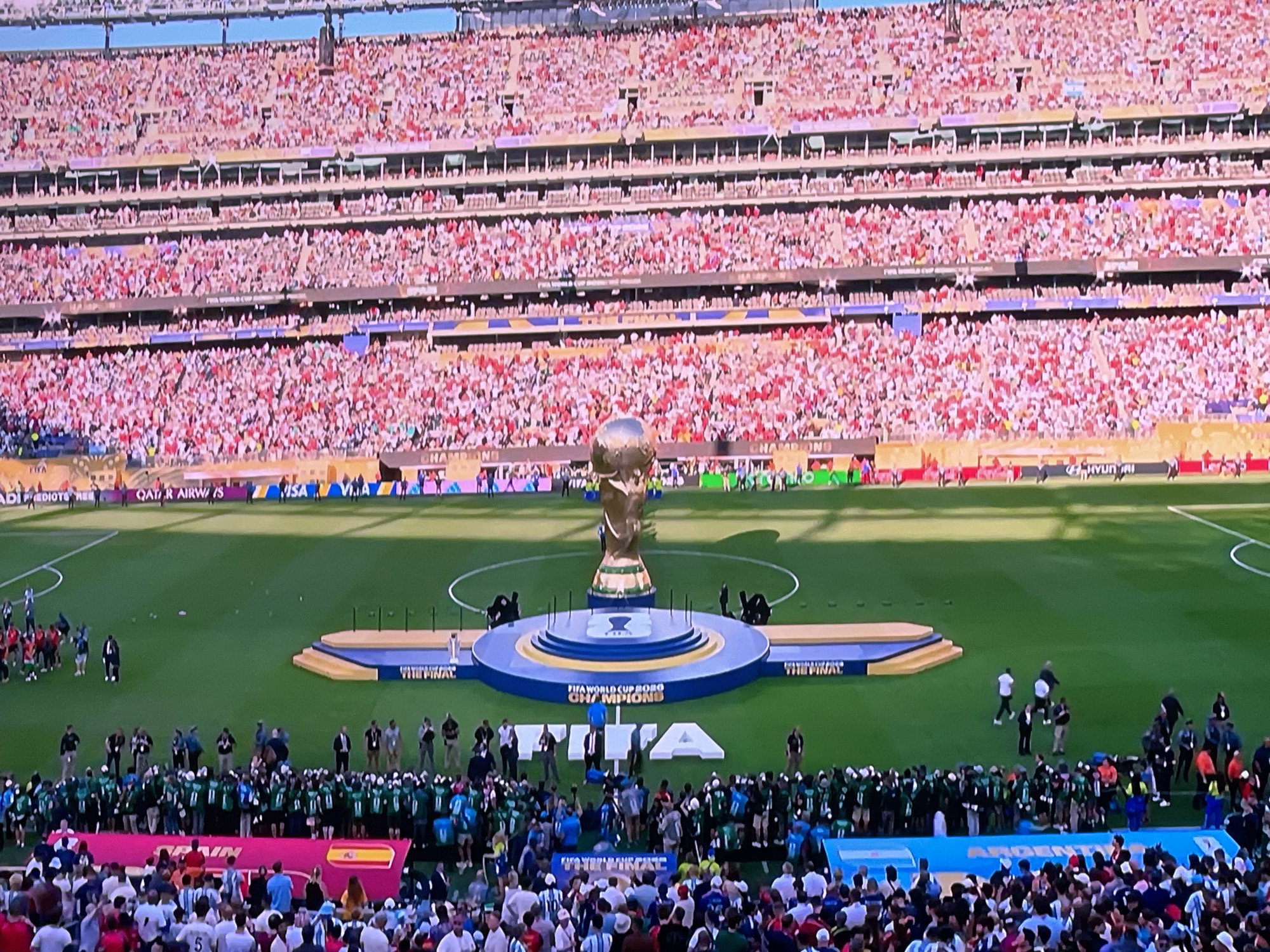 The image depicts a large outdoor stadium filled with spectators, with a soccer field visible in the foreground. A prominent golden trophy, likely the FIFA World Cup, is displayed on a circular platform in the center of the field. A crowd of people, some in green uniforms and others in various attire, are gathered on the field around the trophy. Spectators are seated in the tiered stands that surround the field, with many wearing red and white clothing. The overall scene suggests a major sporting event, possibly the conclusion of a tournament. The image does not contain any information that places it in the city of Stary Oskol, Russia.