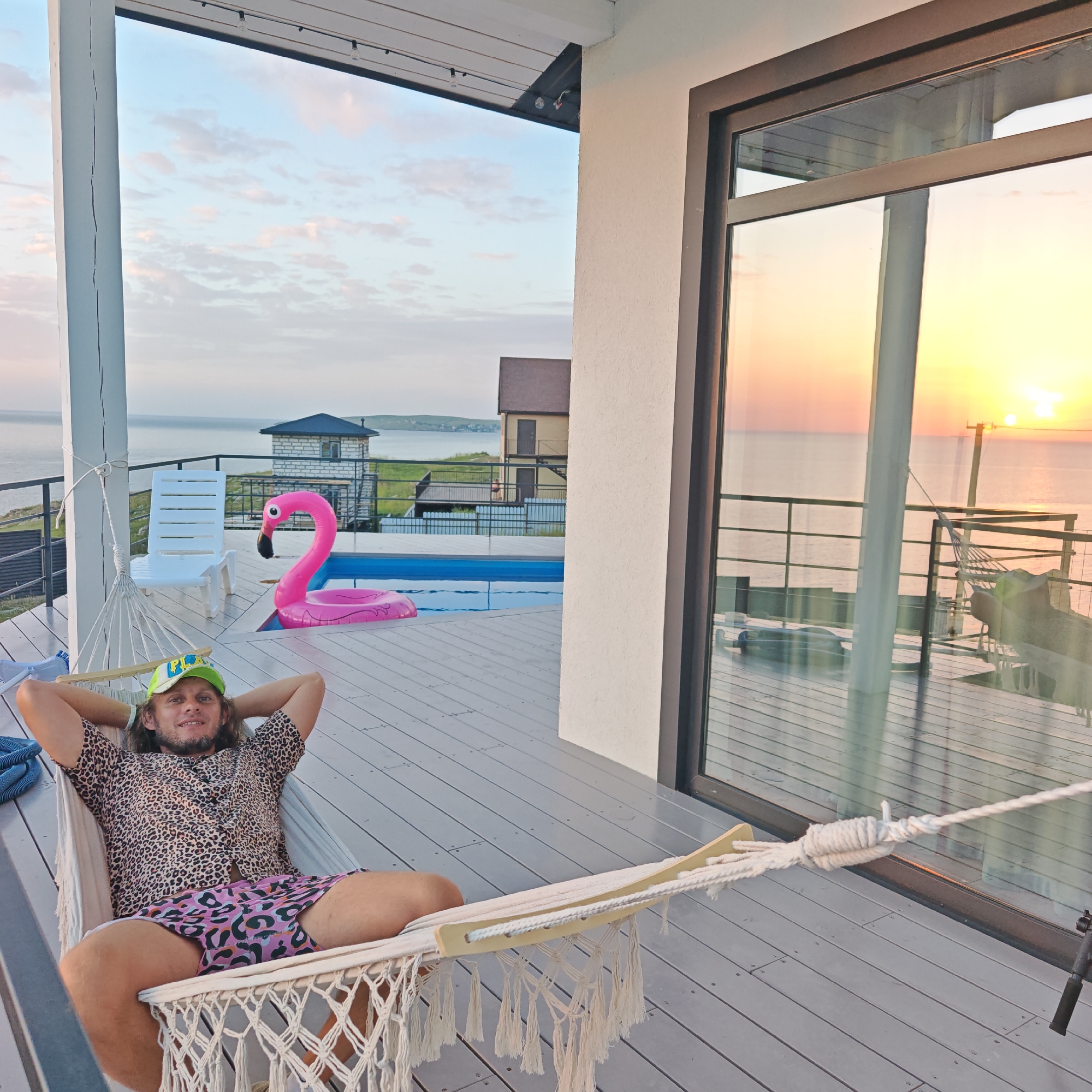 The image shows a man in a hammock on a deck. The man has shoulder-length hair, a beard, and is wearing a leopard-print shirt, pink patterned shorts, and a green baseball cap. He is lying in the hammock with his hands behind his head. Behind him is a pool with a pink flamingo float. A white plastic chair is placed near the pool. Beyond the pool is a body of water and a distant coastline. The setting sun is visible through a large glass door to the right. The location is near the city of Unknown, Unknown, as per the data provided.