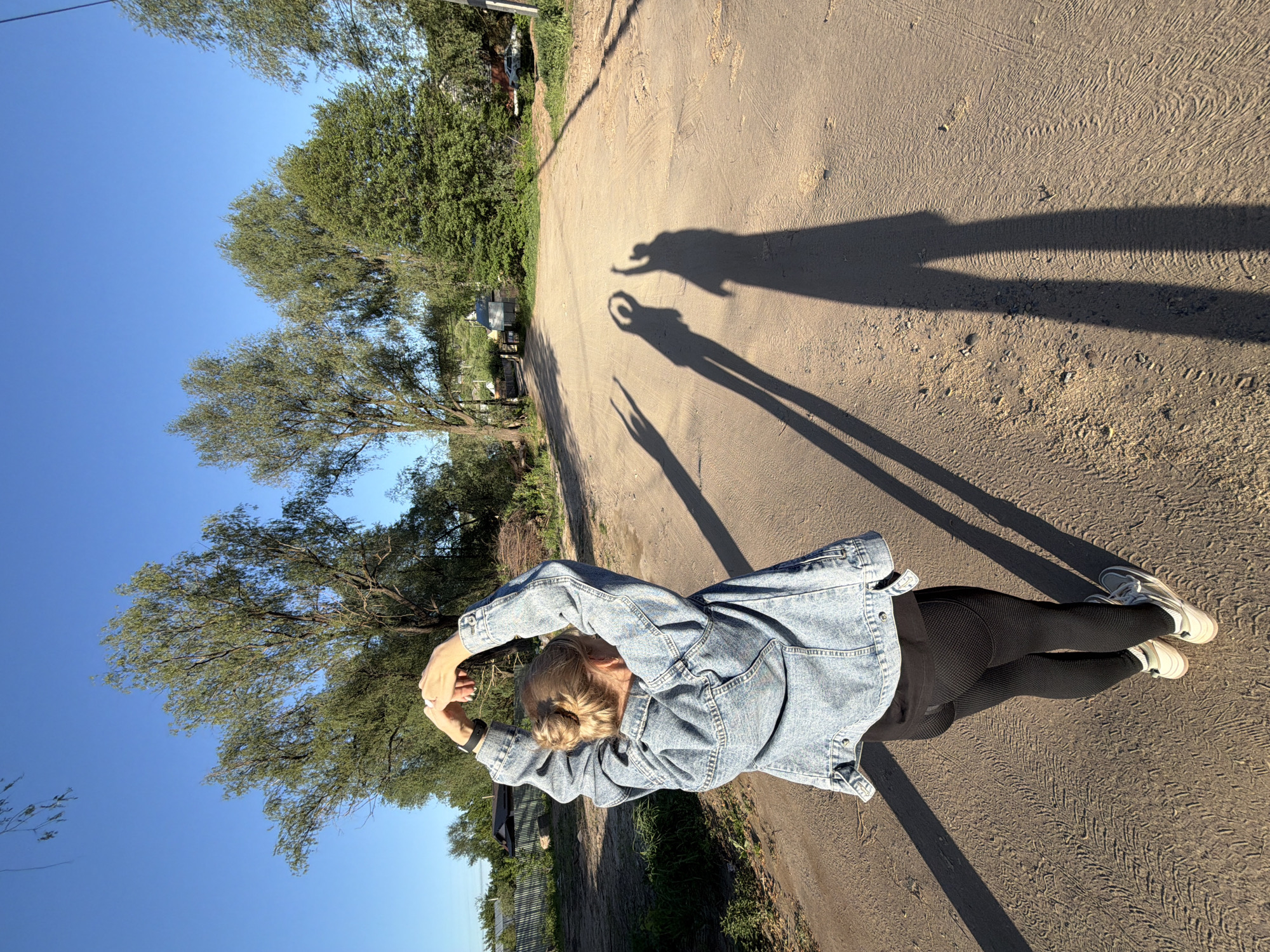 A ground-level, angled shot features a person on a dirt road with trees on the left, likely a road on the outskirts of the city of Chaikovsky, Russia. The person, facing the camera, is wearing a denim jacket, black leggings, and white shoes. Their arms are raised, creating shadow figures on the ground ahead. The area is sunny and likely dry, as seen by the dirt road and shadows.