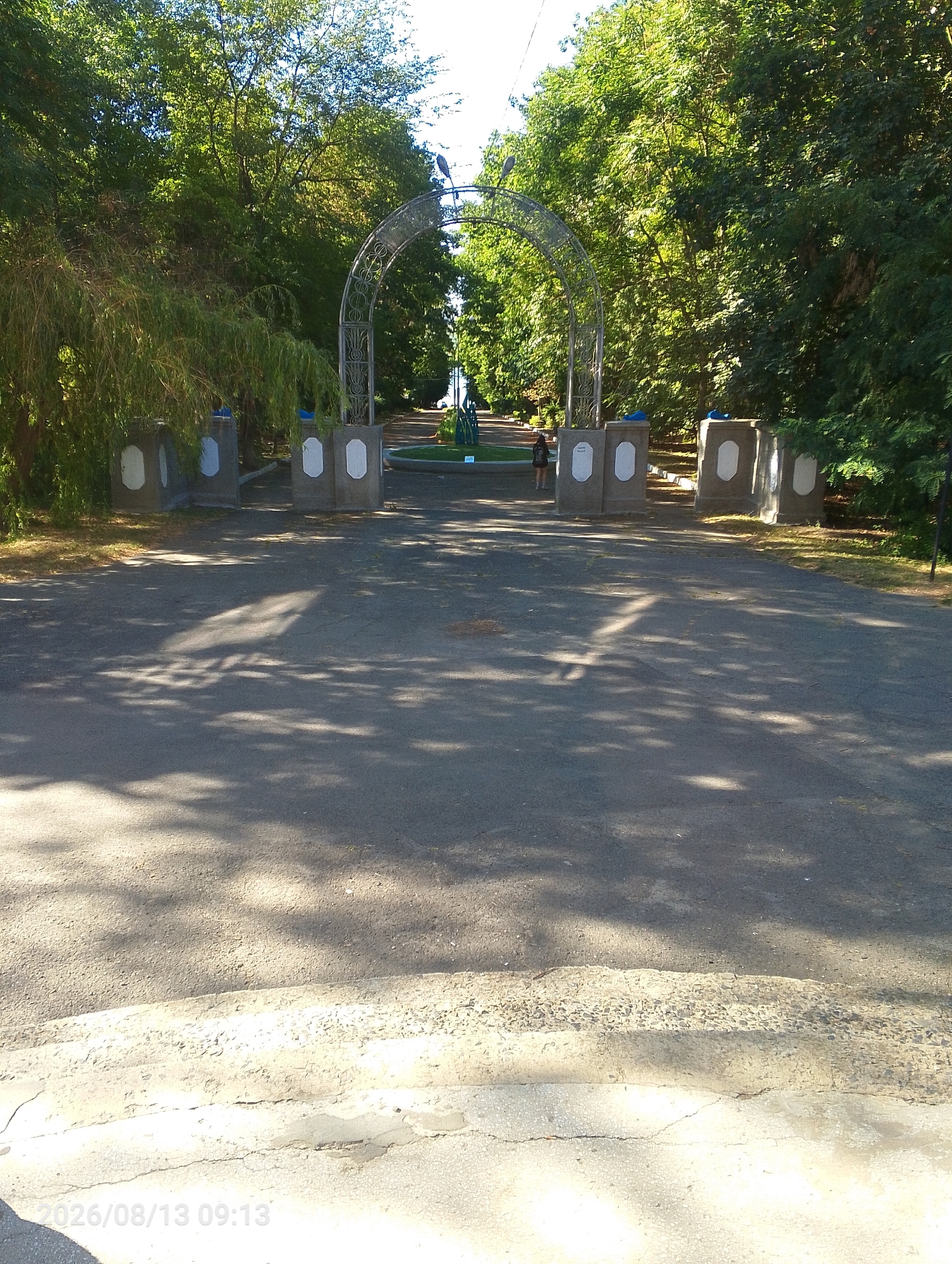 The image shows an entrance to a park in Bahacheve, Ukraine, on a sunny day. The entrance is marked by a metal archway with decorative scrollwork. On either side of the pathway leading through the arch are two concrete pillars topped with decorative elements. Beyond the arch, a paved path continues, lined with trees and greenery, and a circular green space is visible. A person is walking away from the camera towards the archway. The foreground shows a paved surface with dappled sunlight and shadows from the surrounding trees. A watermark at the bottom left corner indicates the date "2026/08/13 09:13".