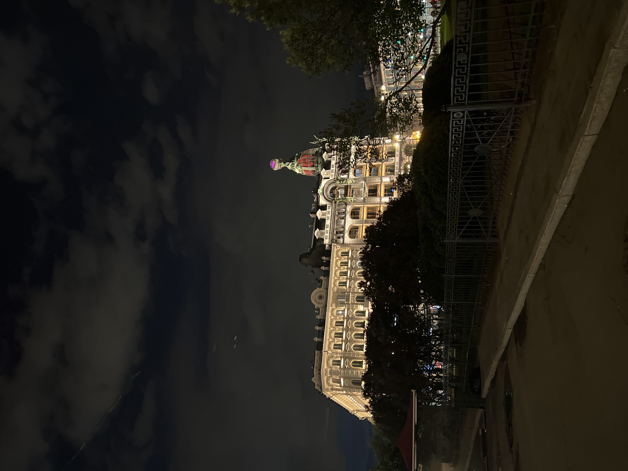 The image shows a nighttime view of the Singer House (also known as the House of Books) in Saint Petersburg, Russia. The building is brightly lit and stands out against the dark sky with scattered clouds. To the right, a wrought iron fence runs along a sidewalk, partially obscured by trees. The scene appears to be taken from street level, looking up at the illuminated building.