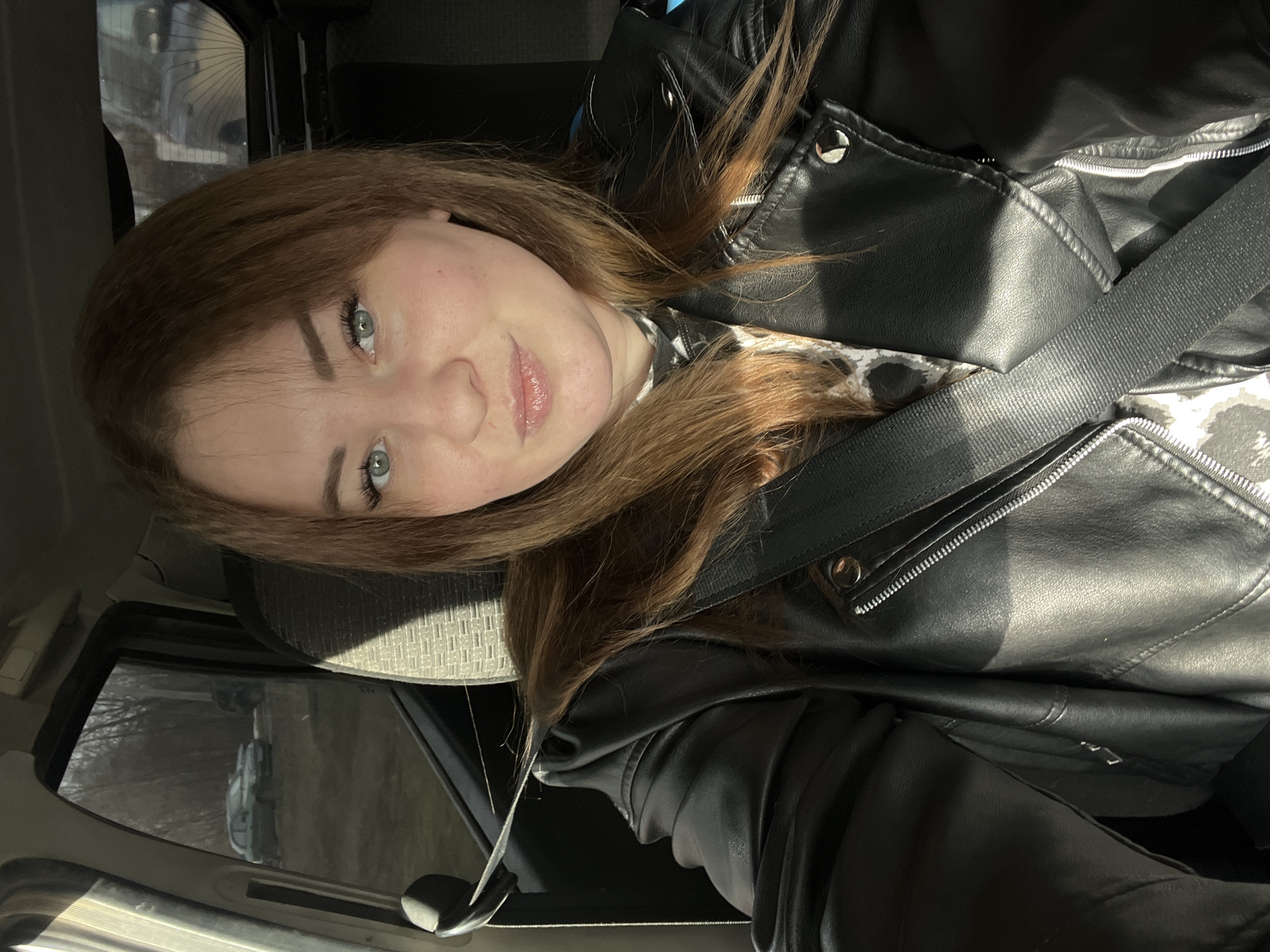 A young woman with shoulder-length reddish-brown hair and light eyes is photographed from a close-up, slightly angled perspective. She is wearing a black leather jacket over a patterned top and is secured with a black seatbelt. She appears to be in a car, based on the interior elements visible, such as the headrest, window, and door. Outside the window, bare trees and a glimpse of another vehicle can be seen, suggesting a possibly rural or suburban environment. It's not possible to confirm the location in Chaikovsky, Russia, solely based on this image.