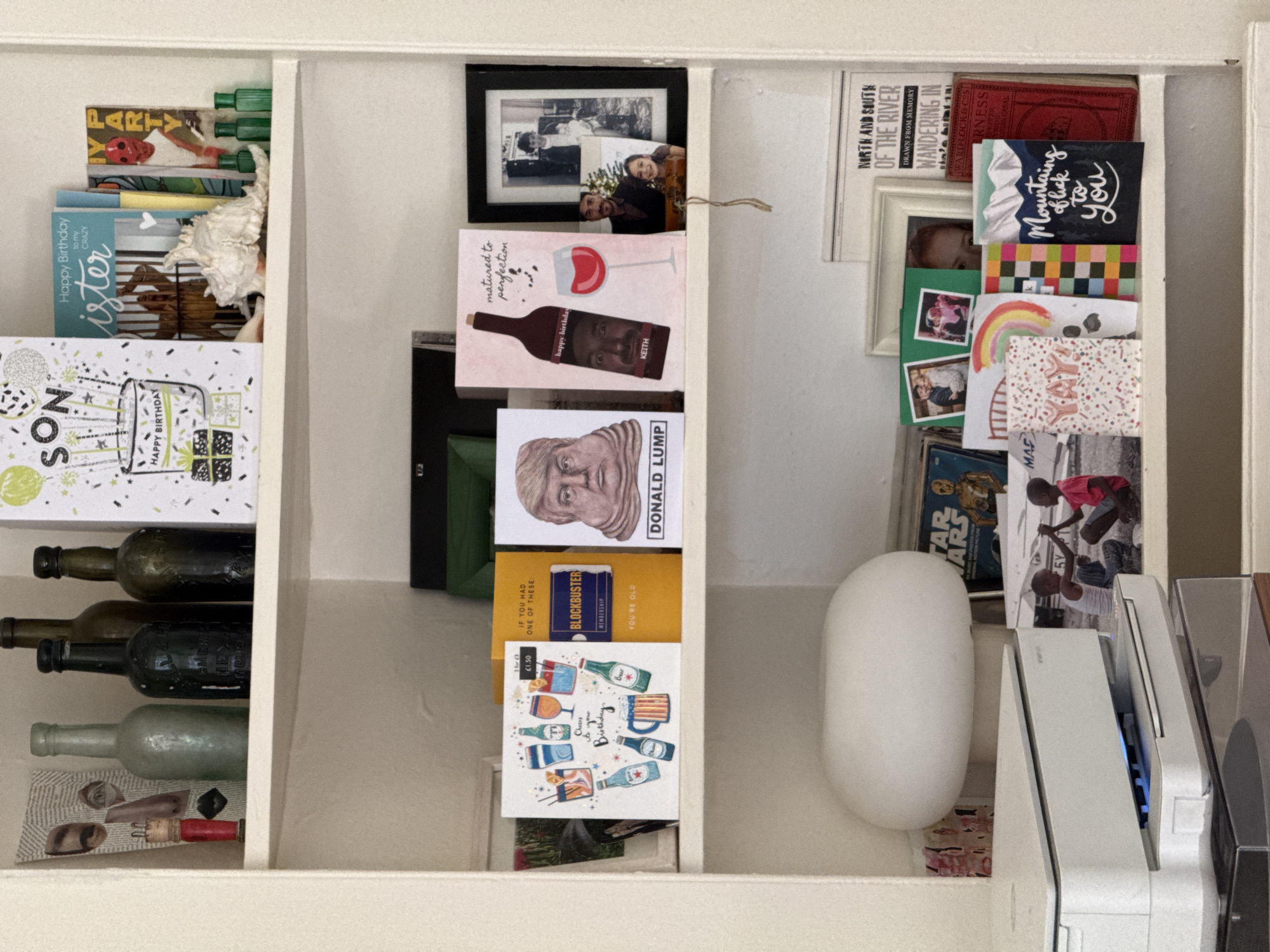 This media file captures a densely packed white shelving unit, displaying a variety of personal items, cards, and decorative objects. The scene is indoors, presenting a collection of cherished memorabilia and greetings.
The left section of the shelf holds green cylindrical objects and a partially visible "PARTY" card. Below these, a white ruffled item is placed next to birthday cards, one for a "CRAZY sister" and another for "SON" with confetti and gifts. The bottom of this section features four vintage-style glass bottles in dark and light green, accompanied by small trinkets including corks and eye-shaped decorative pieces.
The middle section showcases framed photographs, including a black-and-white image of a couple and smaller color photos. A prominent pink birthday card for "KEITH" reads "matured to perfection" and depicts a wine glass and a bottle with a face on the label. Other cards include one with a sculpted head labeled "DONALD LUMP," a nostalgic yellow "BLOCKBUSTER MEMBERSHIP YOU'RE OLD" card, and a "Cheers to your Birthday" card adorned with illustrations of various alcoholic beverages.
The rightmost section contains book spines or covers with titles such as "NORTH AND SOUTH OF THE RIVER" and "WANDERING IN...," alongside a red-bound book. A collection of greeting cards is displayed, featuring designs like mountains with "Mountains thank you," a colorful checkered pattern, a rainbow, and "YAY!" Small photographs of children and adults are interspersed among the cards. Further down, a "STAR WARS" item and a black-and-white photo of two children are visible. The bottom right features a white, oval-shaped object (possibly a smart speaker or lamp) next to white electronic equipment, resembling a printer or scanner, which emits a blue light.
The diverse array of birthday cards suggests a focus on celebrations and personal connections. The objects blend nostalgic elements, like the vintage bottles and Blockbuster card, with more contemporary items, such as the white electronic device. There are no specific cues for time of day or external location.