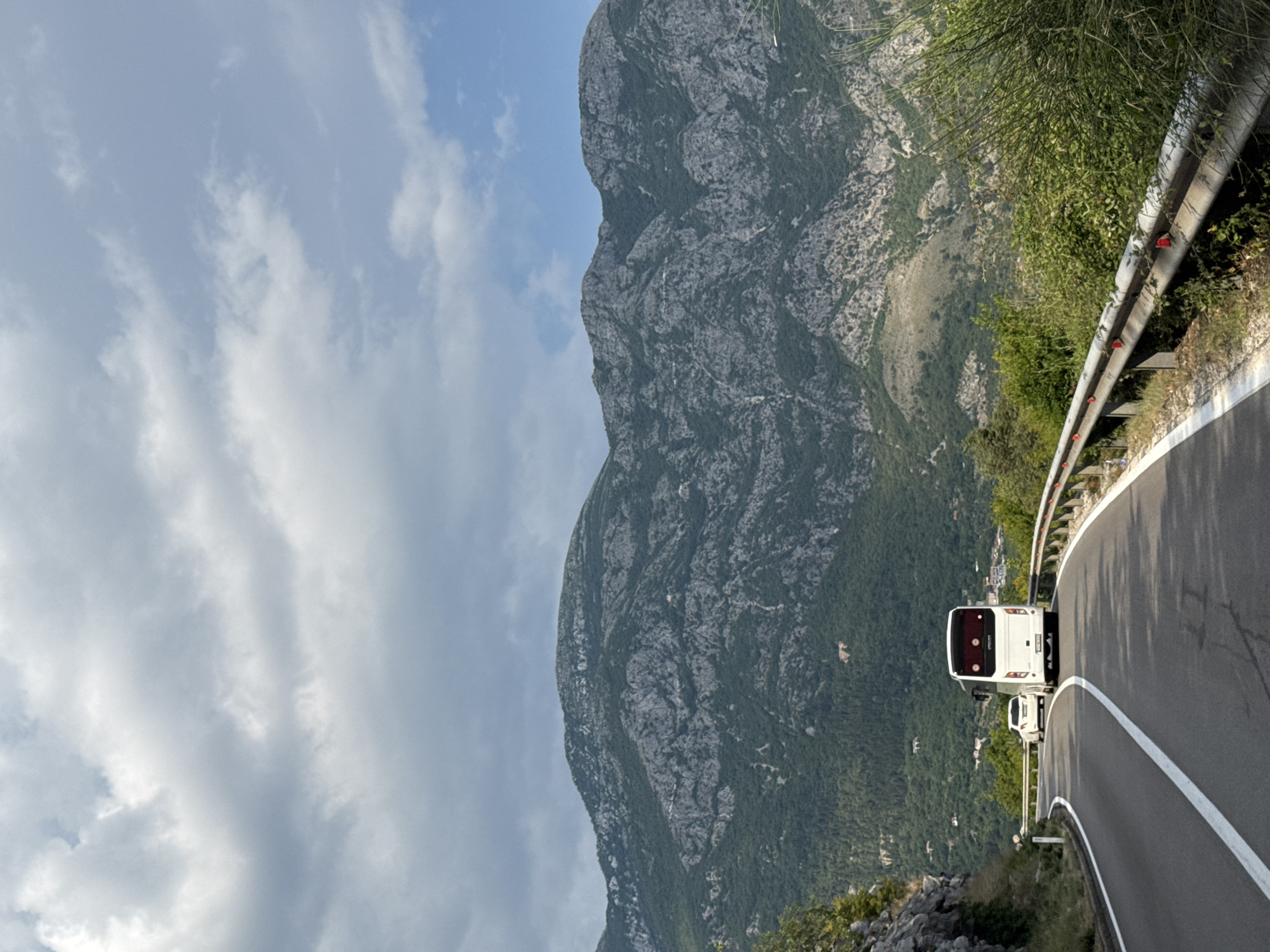 A white bus and a white car drive on a winding road in Strp, Montenegro. The road is paved and has a white line marking the lanes. A metal guardrail runs along the right side of the road. The bus is in the foreground, and the car is behind it. In the background, there are mountains covered in trees. The sky is cloudy.