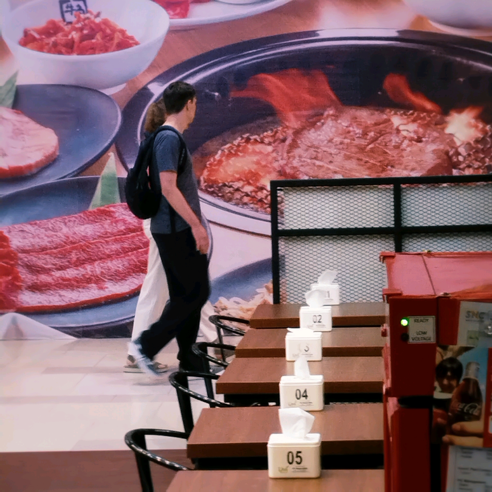 The image depicts an indoor scene, likely a restaurant or food establishment, featuring a large mural of food. In the foreground, there are several tables with tissue box dispensers labeled 02, 03, 04, and 05. A person wearing a dark t-shirt and black pants, with a backpack, is walking from left to right across the frame. Another person, dressed in white pants and a lighter colored top, is walking behind the first person. To the right, a red dispenser unit with a green indicator light labeled "READY" and "LOW VOLTAGE" is visible, along with a Coca-Cola advertisement. The background shows a mural that includes depictions of raw meat dishes, sizzling meat, and a bowl of what appears to be kimchi. The environment suggests a Korean barbecue or similar dining setting. This image was captured in the city of Surabaya, Indonesia.