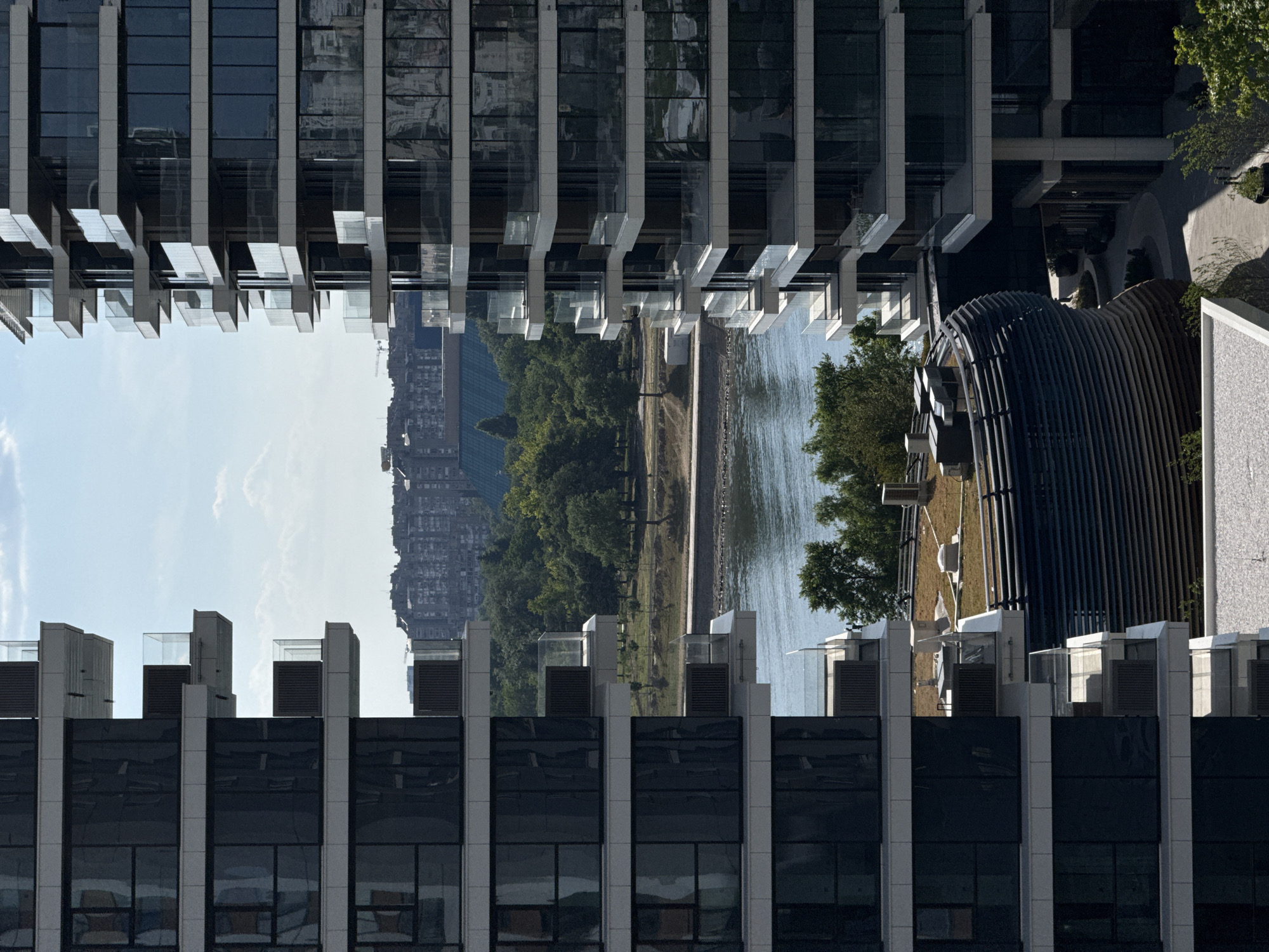 The image shows an urban landscape in Belgrade, Serbia. It is framed by two sections of a modern building with a repeating pattern of windows and vertical structural elements. The view between these building sections reveals a scene with the Sava River, a park with trees and grass, a pathway along the river, and a cluster of buildings in the distance. A distinctive architectural structure with a curved, ribbed exterior, possibly a building's ventilation or a design feature, sits near the right side of the frame. The sky is partially visible in the opening, suggesting a clear or slightly cloudy day. The overall composition has a surreal, Escher-like quality due to the buildings being depicted from different orientations.