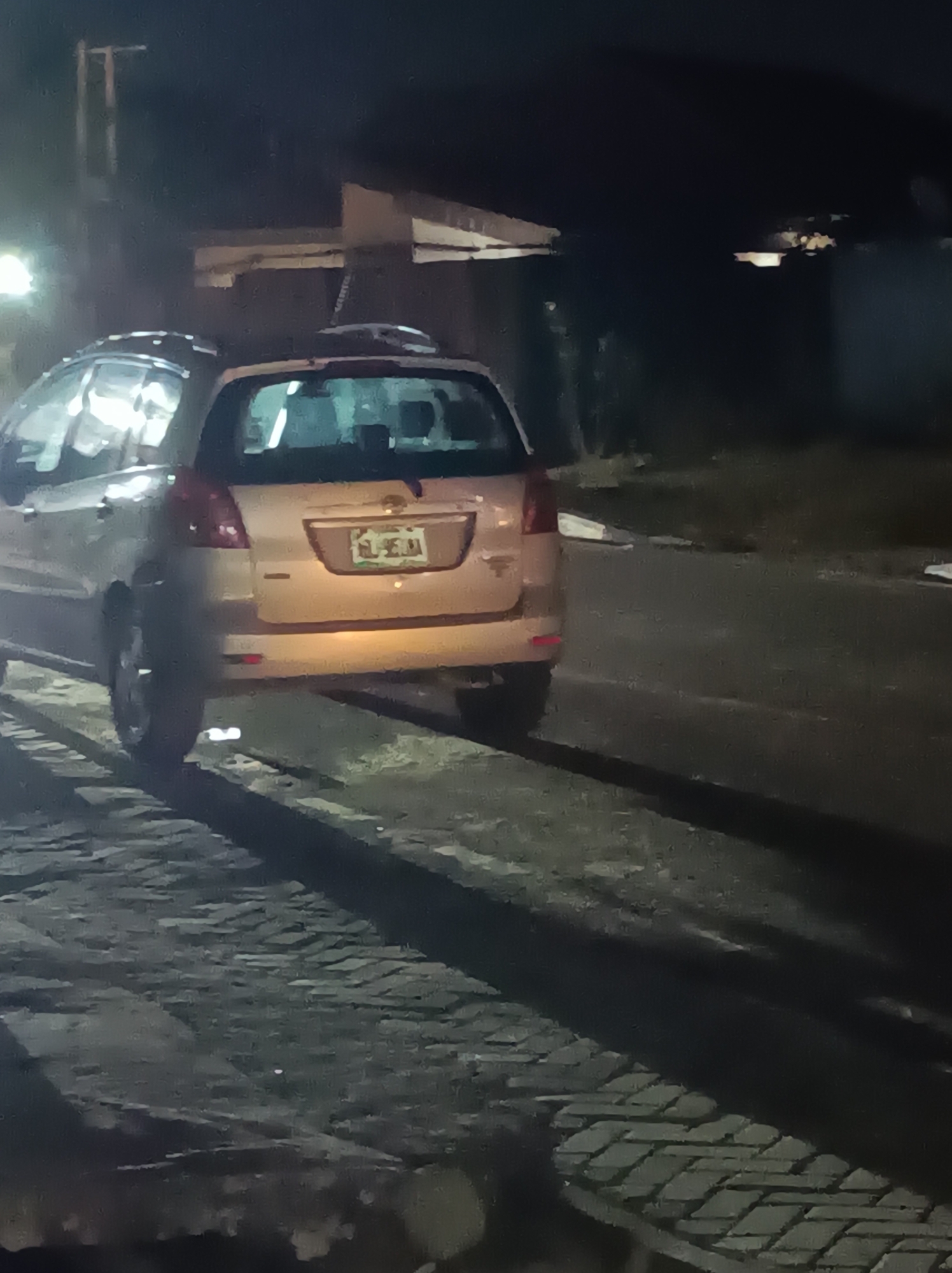 The image shows a light-colored, possibly gold or beige, station wagon or compact SUV viewed from the rear at night in Narayi, Nigeria. The vehicle is positioned on a dark, paved surface, with a patterned brick or cobblestone pathway visible on its left, running alongside the car.
The scene is dimly lit, indicating nighttime. Artificial light sources reflect off the car's body and illuminate the area around its rear, particularly the license plate. The background is dark, with some faint, distant lights visible, and a blurred structural element, possibly a building or part of an overhead structure, in the upper left. No people are visible.
The most prominent detail is the car's license plate, which is illuminated. The top line of the license plate clearly reads "LAGOS", indicating its registration state in Nigeria. The bottom line displays a sequence of characters that are somewhat blurry due to the low light and image quality. It appears to start with "AE", followed by three digits that are difficult to discern precisely but could be "580" or "503", and then ends with "JJ". Thus, the license plate likely reads "LAGOS AE 580 JJ" or "LAGOS AE 503 JJ".
The overall impression is a quiet, nocturnal urban or semi-urban setting, with the car appearing stationary or slowly moving. The image exhibits characteristics of low-light photography, including some noise and softness.