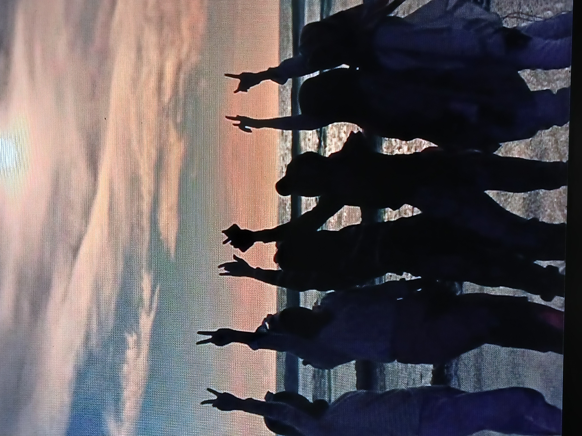 The image depicts a group of people silhouetted against a sky, presumably at sunset, with colors ranging from light blue and white to orange and pink. The people are lined up and appear to be pointing their fingers towards the sunset. The scene is in Juan Griego, Venezuela.