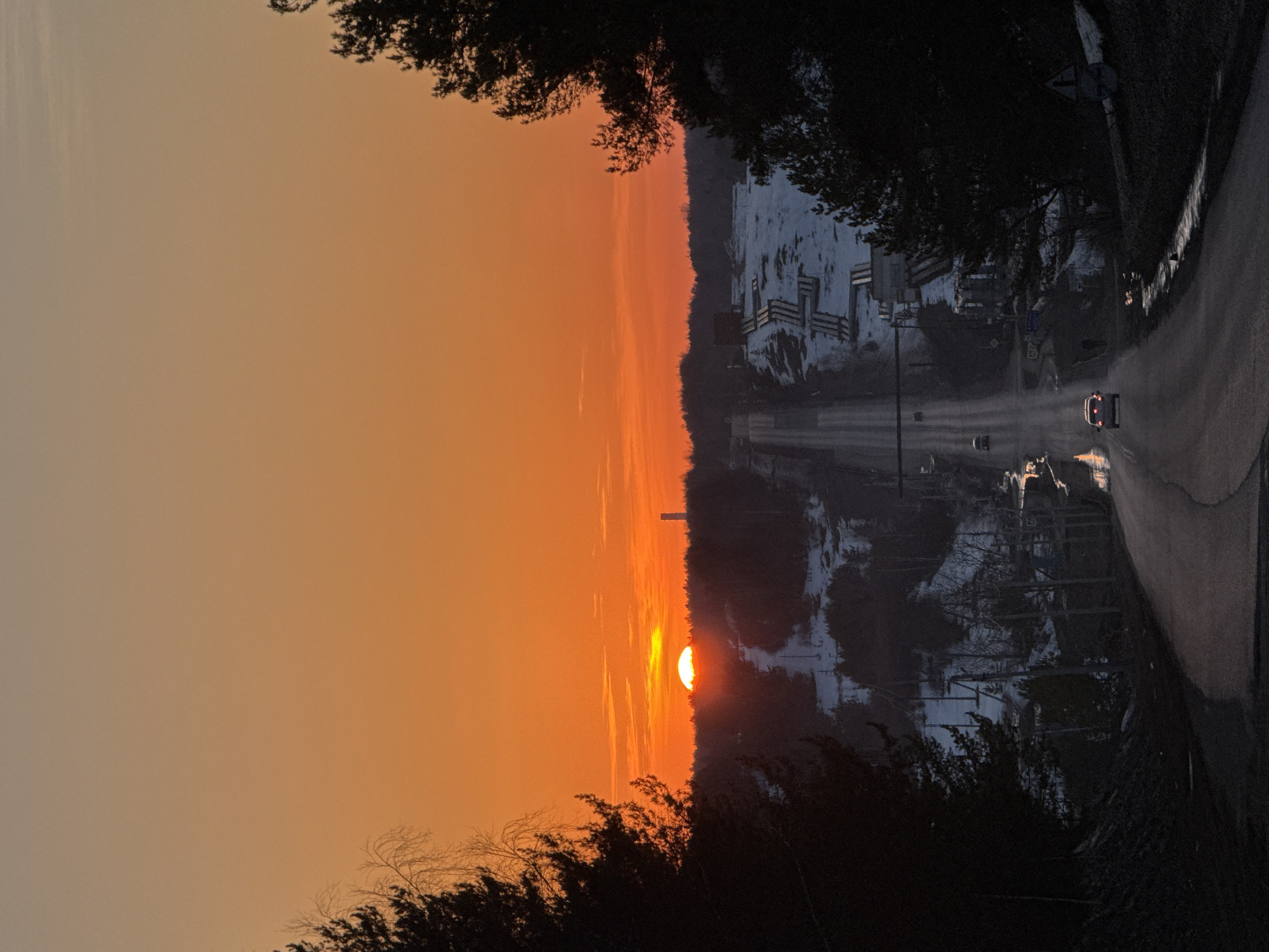 The image depicts a sunset over a road in the city of Прикамский, Russia. The sun is partially obscured by trees and the horizon. The sky is orange and yellow. The road is asphalt and curves to the right. There are streetlights on the side of the road. A car with its headlights on is traveling on the road. Buildings are visible on the left side of the road, some of which are partially covered in snow. Trees line both sides of the road. The season appears to be winter or early spring.