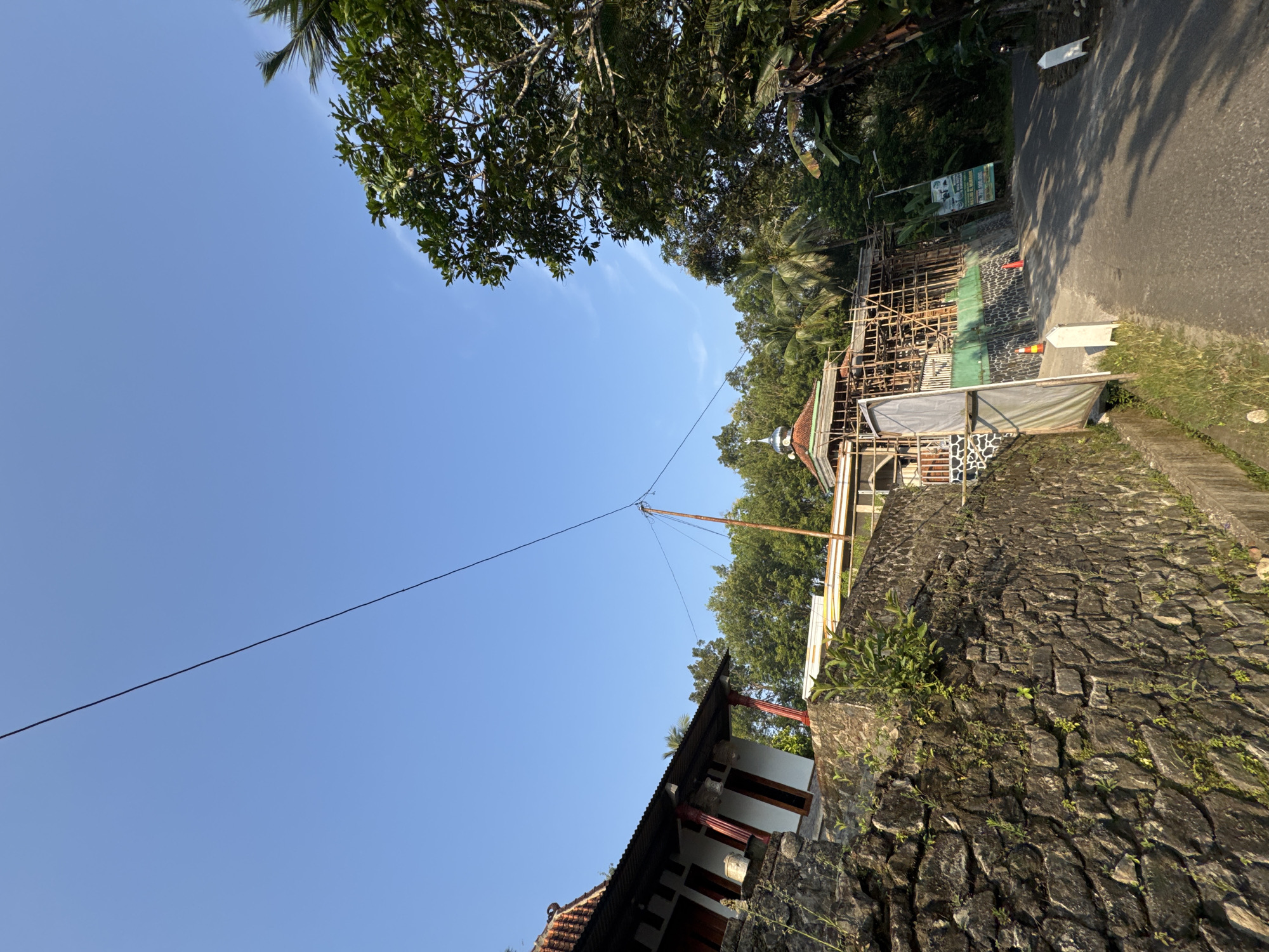 In the city of Alian, Indonesia, a building is undergoing construction, possibly a mosque, as suggested by the dome. The structure is enveloped in scaffolding, indicating ongoing work. A paved road leads up to the building, flanked by a stone wall on the left, covered in moss and vegetation. Lush green trees and palm trees surround the area, creating a tropical ambiance. The sky is clear blue, with a single power line stretching across it, supported by a wooden pole. A white building with a red-tiled roof and ornate columns is visible on the left, situated on top of the stone wall. Several traffic cones are placed near the construction site. A banner with Indonesian text is attached to the fence. The scene is bathed in the warm light of what appears to be late afternoon. There are no people visible in the image.