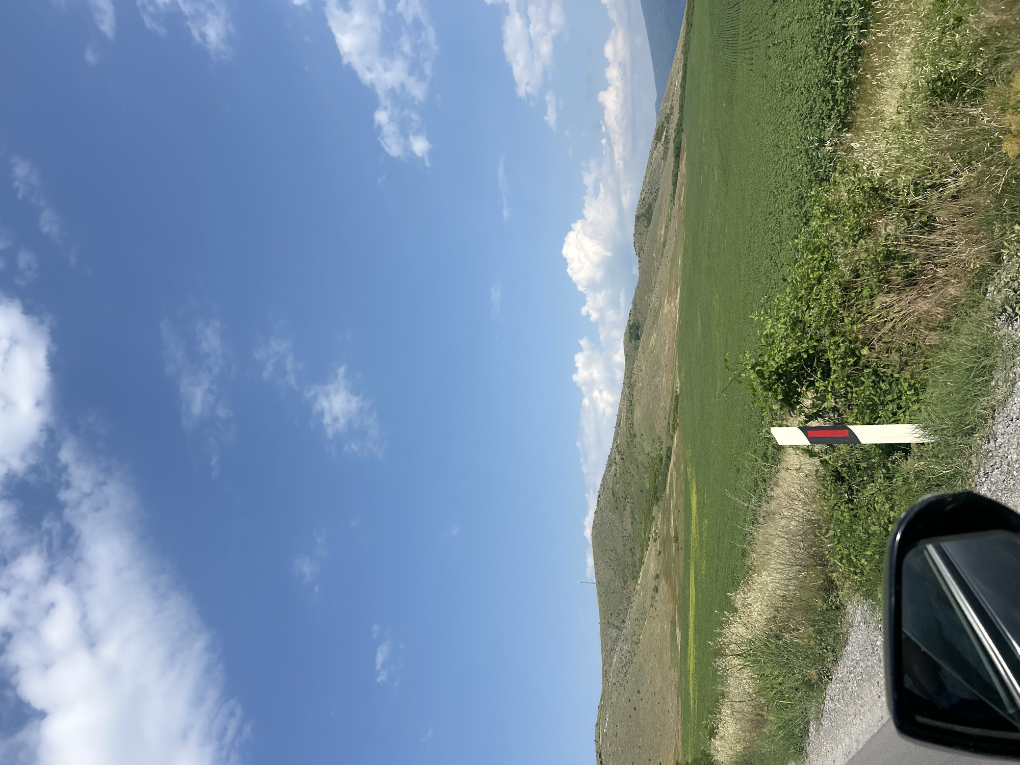 This image depicts a rural landscape in the vicinity of Isperihovo, Bulgaria, under a clear blue sky adorned with scattered white cumulus clouds. In the background, undulating hills covered in dry, light green, and brown vegetation stretch across the horizon. A slender, light-colored tower or mast stands atop the prominent hill in the left-center. Below the hills, agricultural fields are visible; a vast expanse of vibrant green, low-lying row crops dominates the lower midground and extends to the right. Patches of yellowish and greener vegetation are present closer to the base of the hills on the left. The foreground features the edge of a road with a grey gravel shoulder, bordered by a dense strip of wild vegetation composed of dry grasses and green bushy plants. A white rectangular reflector post with a vertical red stripe is positioned within this vegetation, slightly to the right of the lower center. The top portion of a vehicle's side mirror is visible in the bottom-left corner, reflecting internal elements of the vehicle.