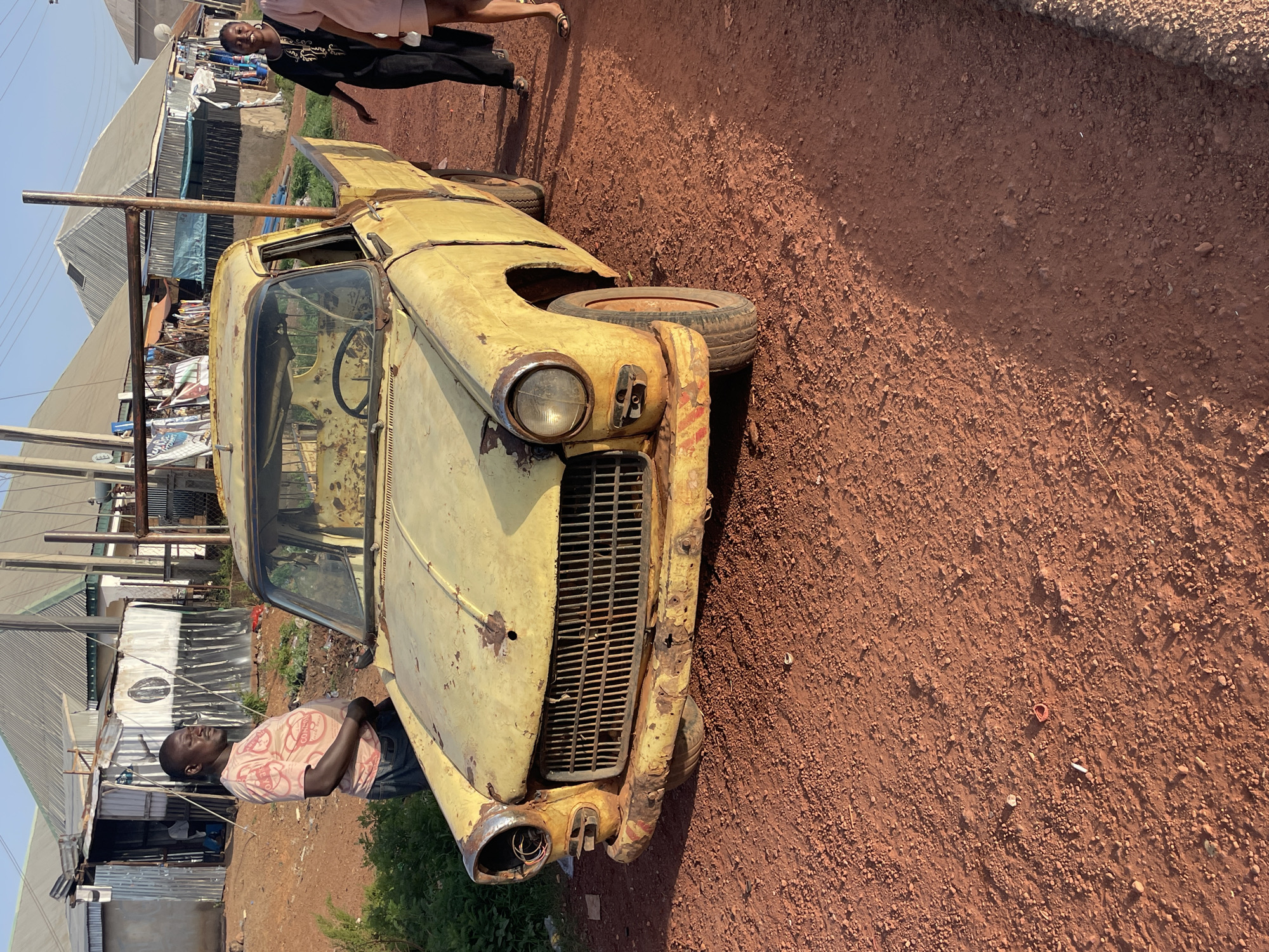 The image shows an old, yellow car in a state of disrepair, with rust and missing parts. The car is located on red, dirt ground, likely a roadside in Durumi, Nigeria, as there are shacks or structures built with corrugated metal sheets in the background. Two individuals are present: one leaning against the car wearing a pink shirt with a faded graphic of the continent of Africa, and another in the background with an open mouth wearing a black shirt and pants, and tan sandals. The scene is in daylight. A metal pole structure is mounted onto the vehicle.