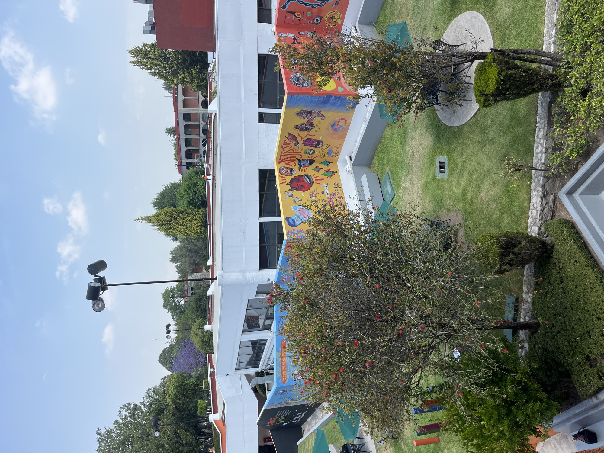 The image captures a vibrant outdoor scene on a clear, sunny day, likely a campus or courtyard in San Andrés Cholula, Mexico. The central focus is a white multi-story building adorned with a large, colorful mural that spans a significant portion of its facade.
The mural itself is a striking piece of public art, divided into several distinct sections with varying color palettes and themes. The uppermost visible section has an orange-red background featuring cartoon-like figures, abstract shapes, and what appears to be a skateboarder. Visible text here includes "PASTEL". Below this, a section with a bright yellow background displays more characters, including a skull, figures playing musical instruments, and stylistic lettering that reads "CAJA", "SKATE", "MUSIC", and "ROCK". A lower section features a blue background with further graphic elements.
In front of the mural-decorated building, there is a meticulously maintained green lawn. Scattered across the lawn are several trees and bushes, including a prominent tree in the foreground with small red flowers or berries. To the right, a circular concrete patio holds dark metal outdoor tables and chairs, suggesting a seating or gathering area. Another, smaller patio is partially visible further to the left. The landscaping also includes trimmed green hedges and low stone walls.
In the background, beyond the main building and trees, other structures with red-tiled roofs and arched corridors are visible, consistent with Mexican architectural styles. A tall light pole with multiple lamps stands on the left side, reaching towards the clear blue sky, which has a few scattered white clouds. Another tree with distinctive purple blooms, likely a Jacaranda, adds a touch of color to the background foliage on the left. No people are visible, and the scene appears peaceful and well-kept.