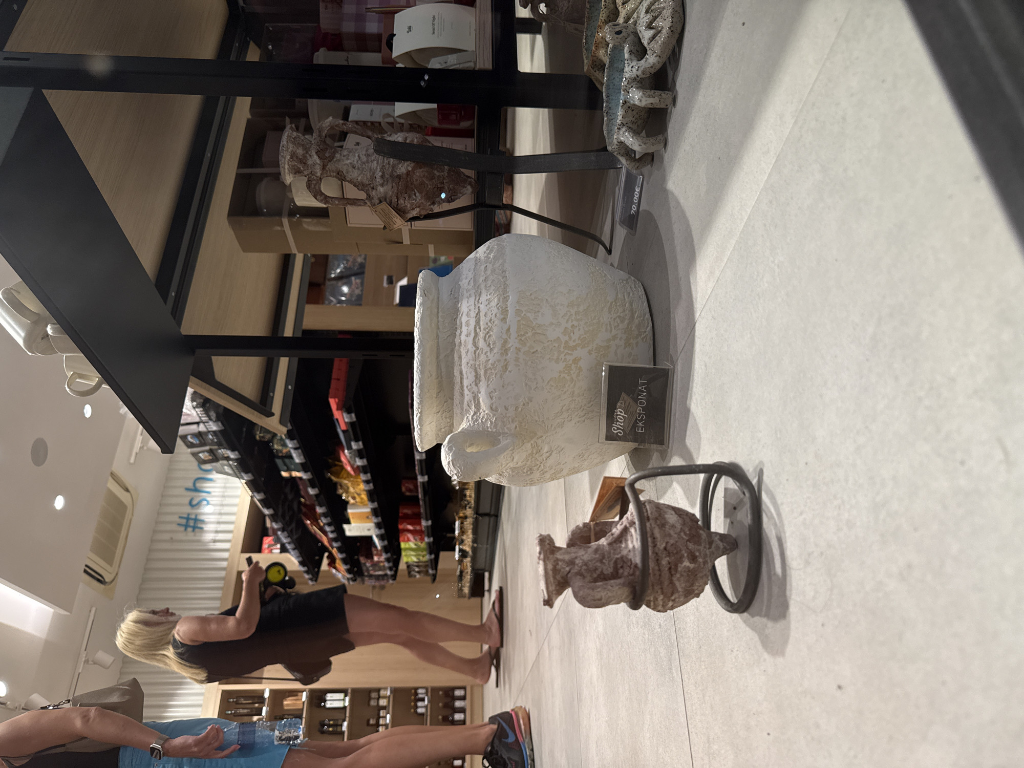 The image shows the interior of a shop, likely a souvenir or gift shop, with a focus on decorative items. In the foreground, a large, textured white ceramic pot is prominently displayed. To its left, a smaller, antique-looking amphora-shaped vase sits on a metal stand. A price tag reading "70.00€" is visible on the floor in front of a decorative crab figurine. In the background, shelves are stocked with various products, including bottles and packaged goods. Two women are partially visible. One woman, with blonde hair and wearing a black dress, is looking at the shelves. Another woman, wearing blue shorts, is also in the background. The shop's floor is tiled. The overall impression is of a well-lit retail environment with an assortment of items for sale. Based on the presence of the price tag in Euros, the shop is likely in the European Union or a country that uses the Euro as currency. However, without more specific visual cues, pinpointing the exact city and country, such as Budva, Montenegro, is not possible from the image alone.
