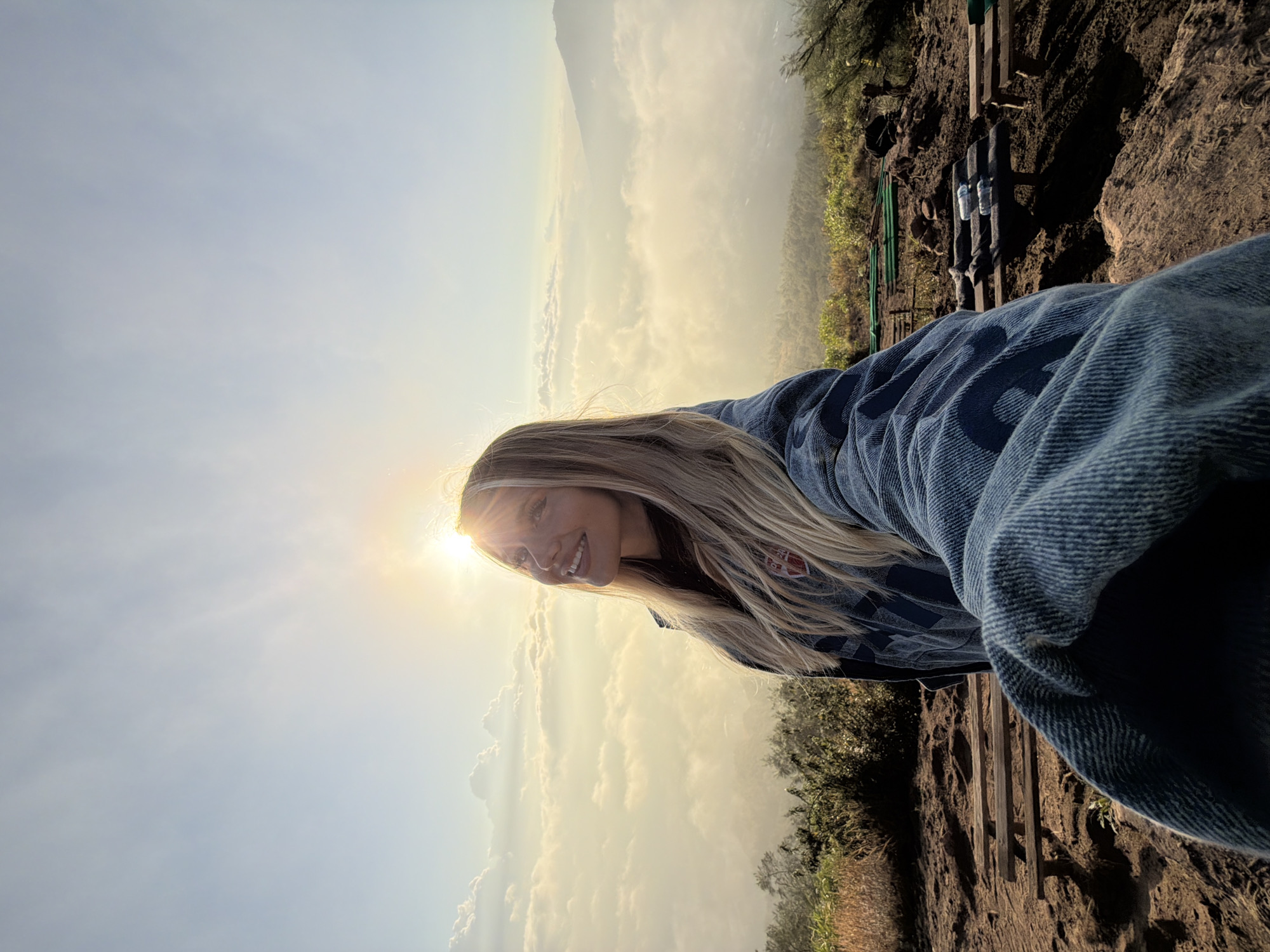 A woman with blonde hair is taking a selfie from a high vantage point overlooking a valley filled with clouds, with mountains in the background. The sun is low in the sky, casting a warm glow over the scene. The woman is wearing a denim jacket and a dark shirt. In the foreground, there is a dirt path with a wooden picnic table and benches. The location is Kintamani, Indonesia.