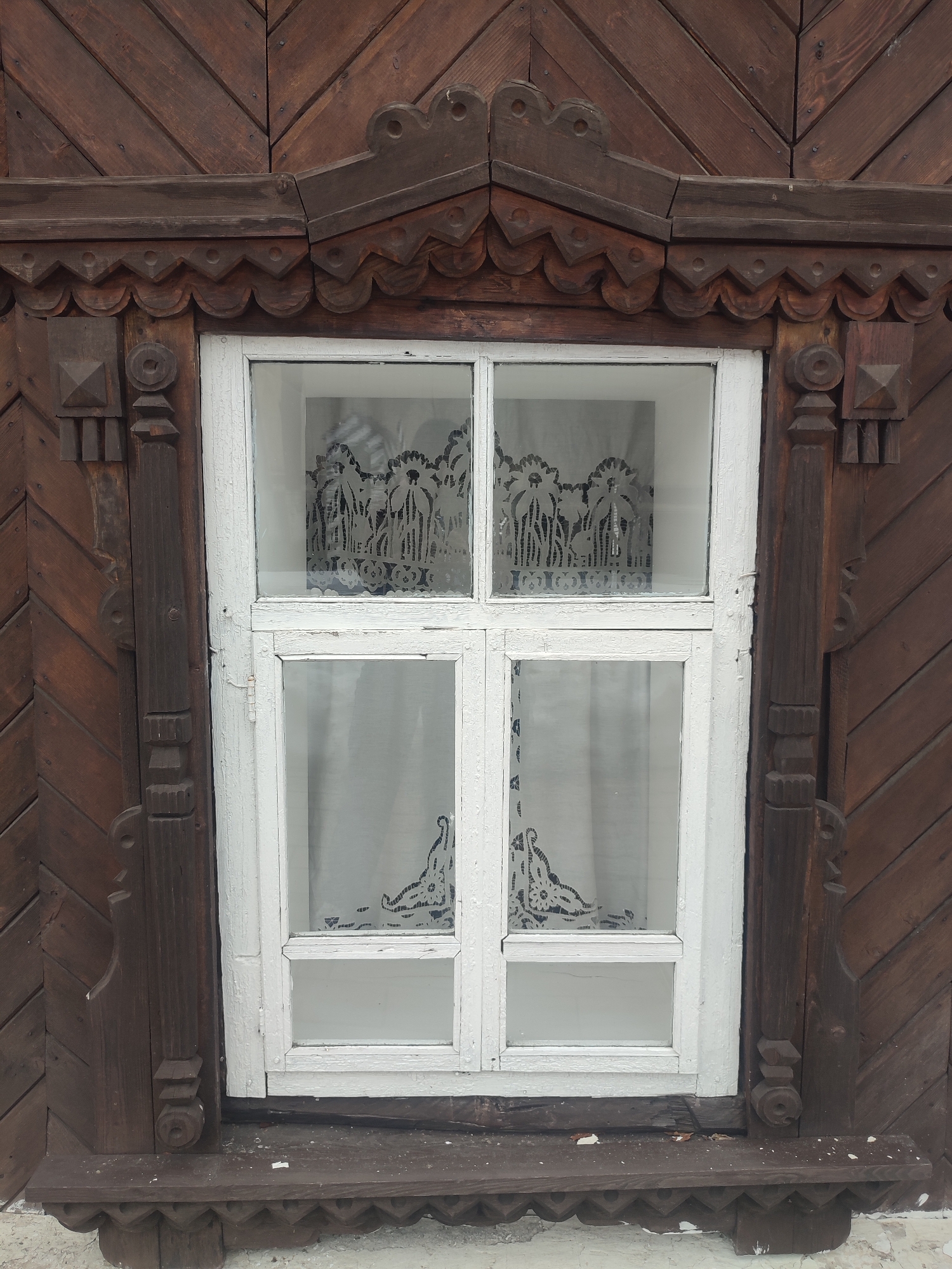 A close-up shot showcases a wooden window frame in Sarapul, Russia. The frame, painted white, is divided into four panes, through which a lace curtain with a floral pattern is visible. The wooden exterior surrounding the window is dark brown, featuring intricate carvings and decorative elements along the top, sides, and bottom of the window frame. The overall style suggests traditional Russian folk architecture.
