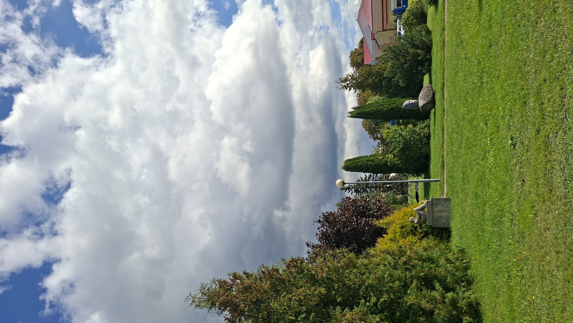 This image captures a portion of a park or green space in Myadzyel, Belarus, on a day with dynamic cloud cover. The foreground is dominated by a lush, green lawn with scattered small yellow flowers. A pathway or mown strip of grass runs horizontally across the mid-ground, separating the lawn from a cluster of trees and decorative elements.
To the left, a large, dense evergreen tree with hints of browning foliage stands prominently. Adjacent to it, a smaller, bright yellow-green shrub is visible. Further into the mid-ground, a stone pedestal supports a sculpture of a reclining human figure.
Behind the pedestal, a tall, slender lamppost with a spherical light fixture rises. Several tall, conical evergreen trees are spaced throughout the scene. To the right of the lamppost, a large, rough-hewn boulder is topped with a sculpted human head, facing towards the left. Behind these elements, a building with a red roof and light-colored walls is partially visible, with a white car parked in front of it.
The background is filled with a mix of blue sky and dramatic, grey and white clouds, suggesting a partly cloudy or changeable weather condition. The overall impression is that of an urban park setting with artistic installations and well-maintained landscaping.