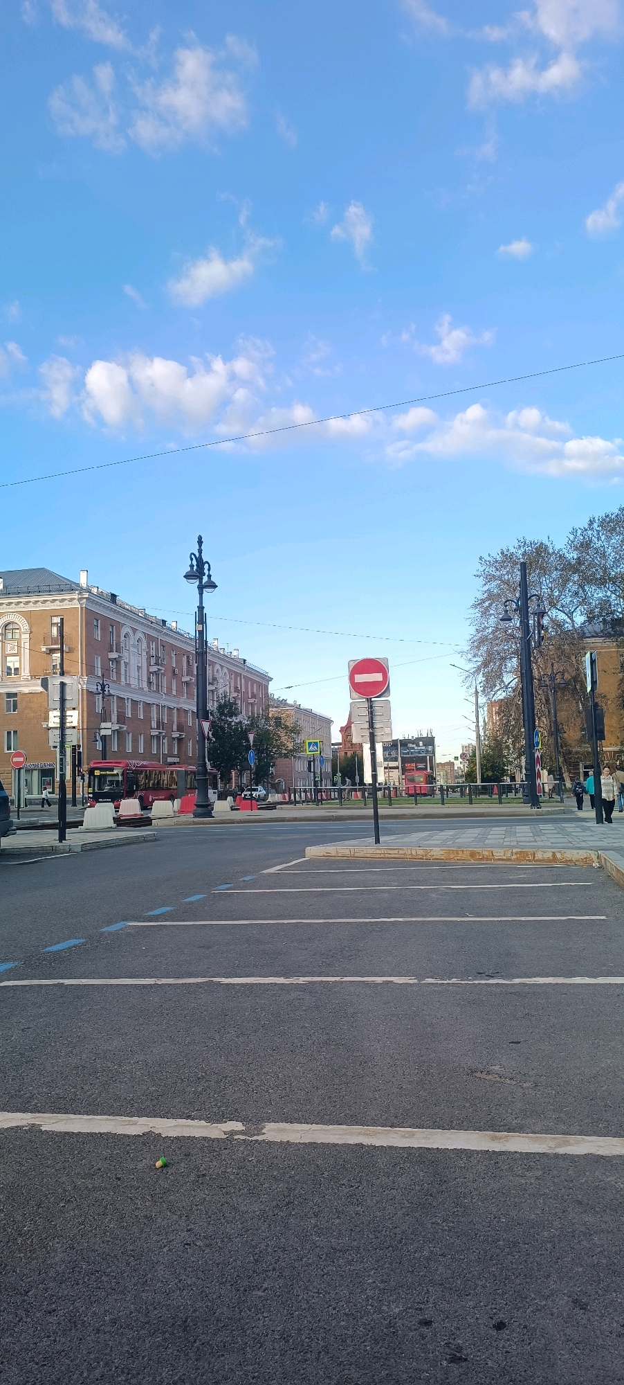 The image depicts a city street scene in Perm, Russia, on a clear day with scattered clouds. In the foreground, there is an asphalt road with white lane markings. A small, green and yellow object, possibly a toy or debris, lies on the road surface.
In the midground, a multi-lane road is visible, with several vehicles including a red bus. Traffic signs are present, including a red circular "no entry" sign. Buildings line the street, characterized by classical architectural styles with multiple windows and decorative elements. A tall street lamp with ornate detailing stands prominently. Trees with sparse foliage are also visible, suggesting it might be autumn.
In the background, more buildings and trees can be seen, extending into the distance. People are visible walking on the sidewalk and near the vehicles, indicating urban activity. The overall impression is that of a public thoroughfare in a built-up area.
