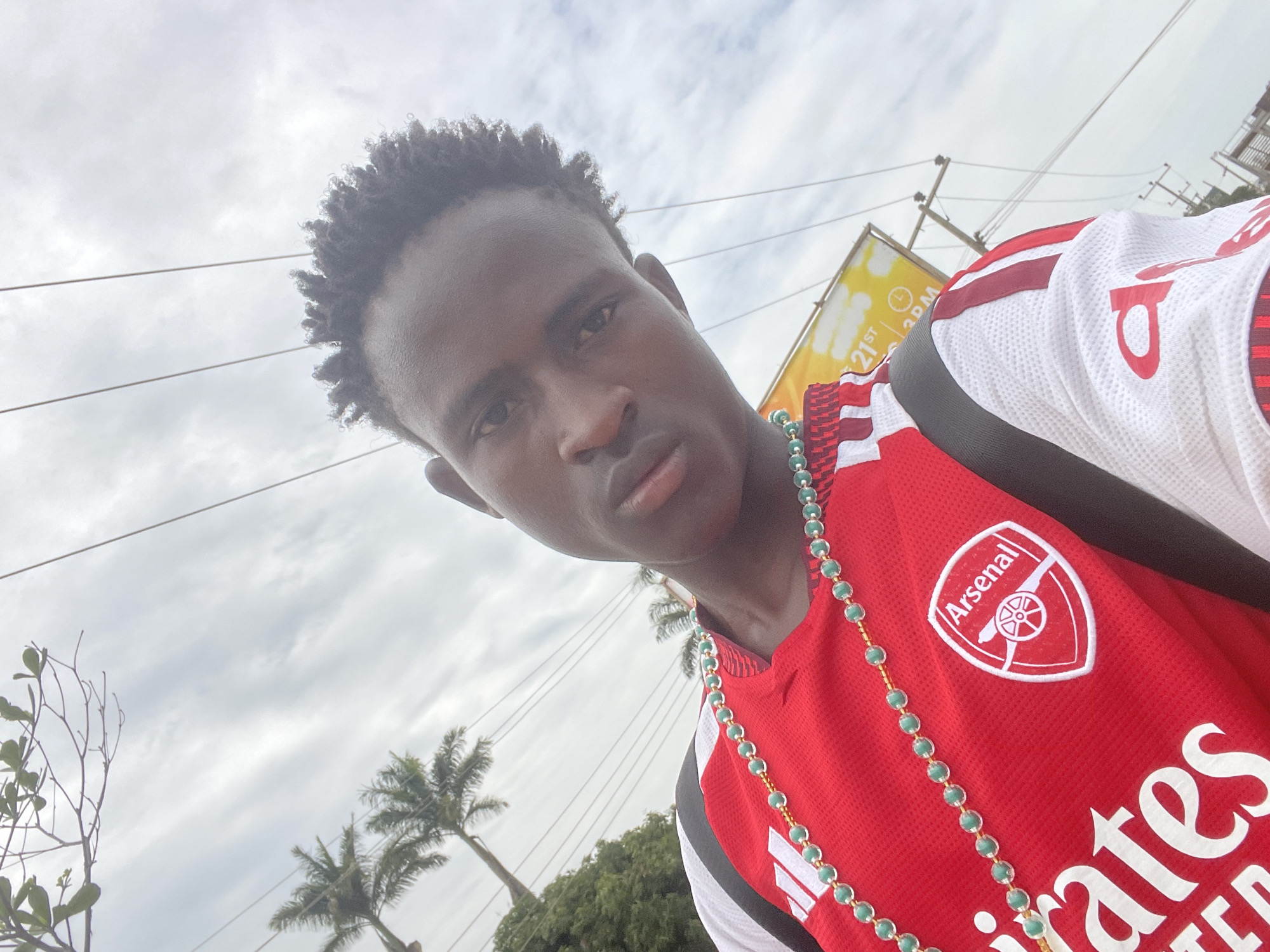 In a close-up, upward-angled selfie, a young Black male is pictured outdoors against a cloudy sky, with palm trees visible in the background. He is wearing a red and white Arsenal football jersey, adorned with a green beaded necklace. The jersey prominently displays the Arsenal logo on the left chest and white Adidas branding on the right. The man's hair is styled in short, dark dreadlocks. Behind him, a yellow sign with text, including "21ST" and "3PM," is partially visible. Electrical wires crisscross the sky above him. The scene suggests an outdoor setting in a city, possibly Jos, Nigeria, given the presence of palm trees and electrical infrastructure.