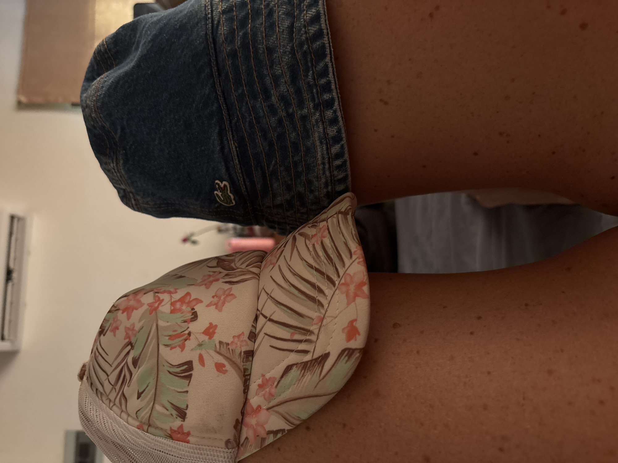 The image shows two caps, one denim and the other patterned with floral and leafy designs, resting on a person's shoulders or upper arms. The denim cap has a small green Lacoste logo. The person's skin tone is tan with visible freckles. The background is slightly blurred and includes parts of a wall and furniture. The image was not taken in บ้านบางรักษ์, Thailand.