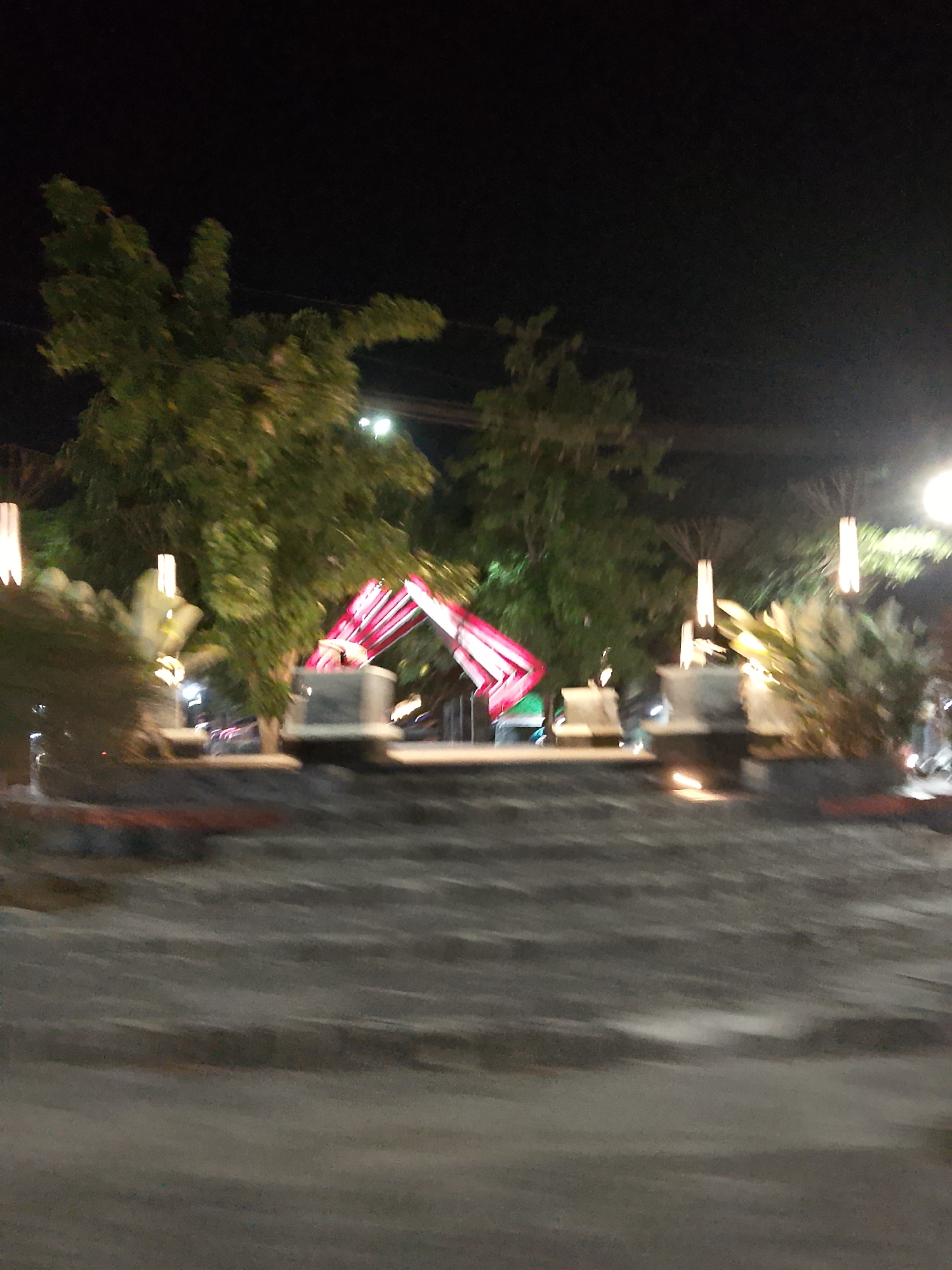 The image depicts an outdoor scene at night, likely a public space or park in Balun, Indonesia. The foreground is dominated by textured gray stairs or a tiered pathway, suggesting an urban or landscaped area. Beyond the stairs, there are several elements that indicate a lively atmosphere:
In the mid-ground, a prominent structure with bright pink and white lights is visible, possibly an art installation or decorative feature. Two individuals are faintly discernible near this structure. Surrounding this area are various plants and trees, some of which are illuminated by soft, warm lighting from vertical fixtures that resemble lanterns or lamps.
In the background, more trees are visible, and a street light with a bright glow is present in the upper right corner. The overall impression is that of a well-lit recreational area or a place of gathering, illuminated for nighttime use. The blurriness of the image suggests it was taken with a low light setting or during movement.