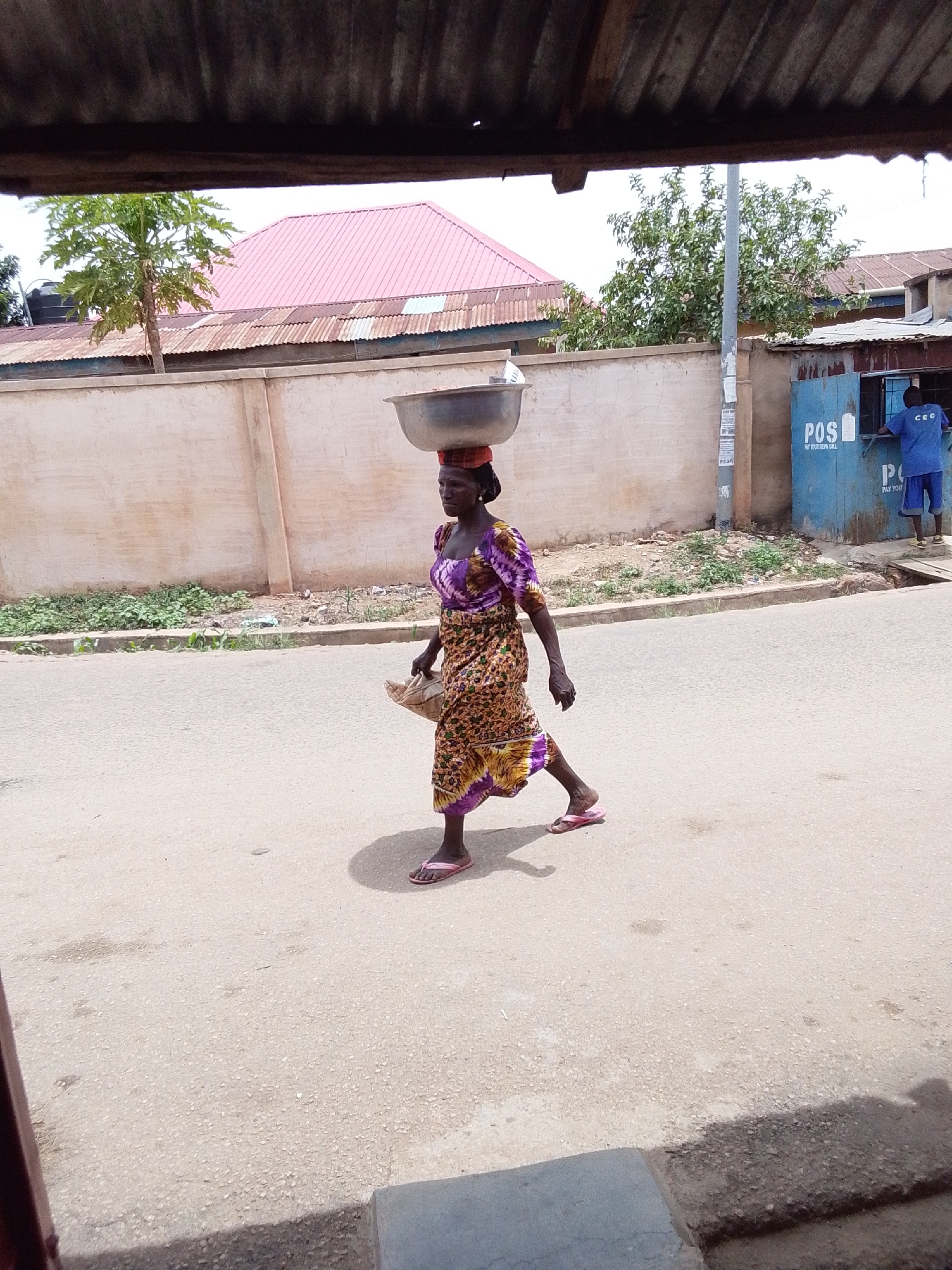 A woman is walking on a street in Jos, Nigeria. She is carrying a large metal bowl on her head and a brown bag in her left hand. She is wearing a colorful dress and pink sandals. The street is paved and there is a building in the background with a pink roof. There is also a POS pay your view will kiosk to the right of the woman. A man in a blue uniform is standing inside of the kiosk.